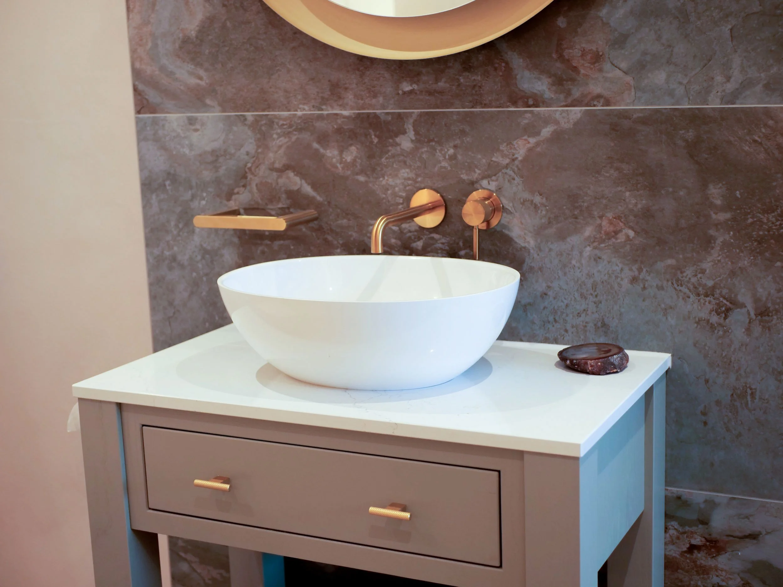 White vessel sink on a modern beige bathroom vanity with gold hardware, bronze faucet, beige countertop, a dark brown soap dish, and a circular mirror above. The wall behind is covered in brown and reddish marble tiles.