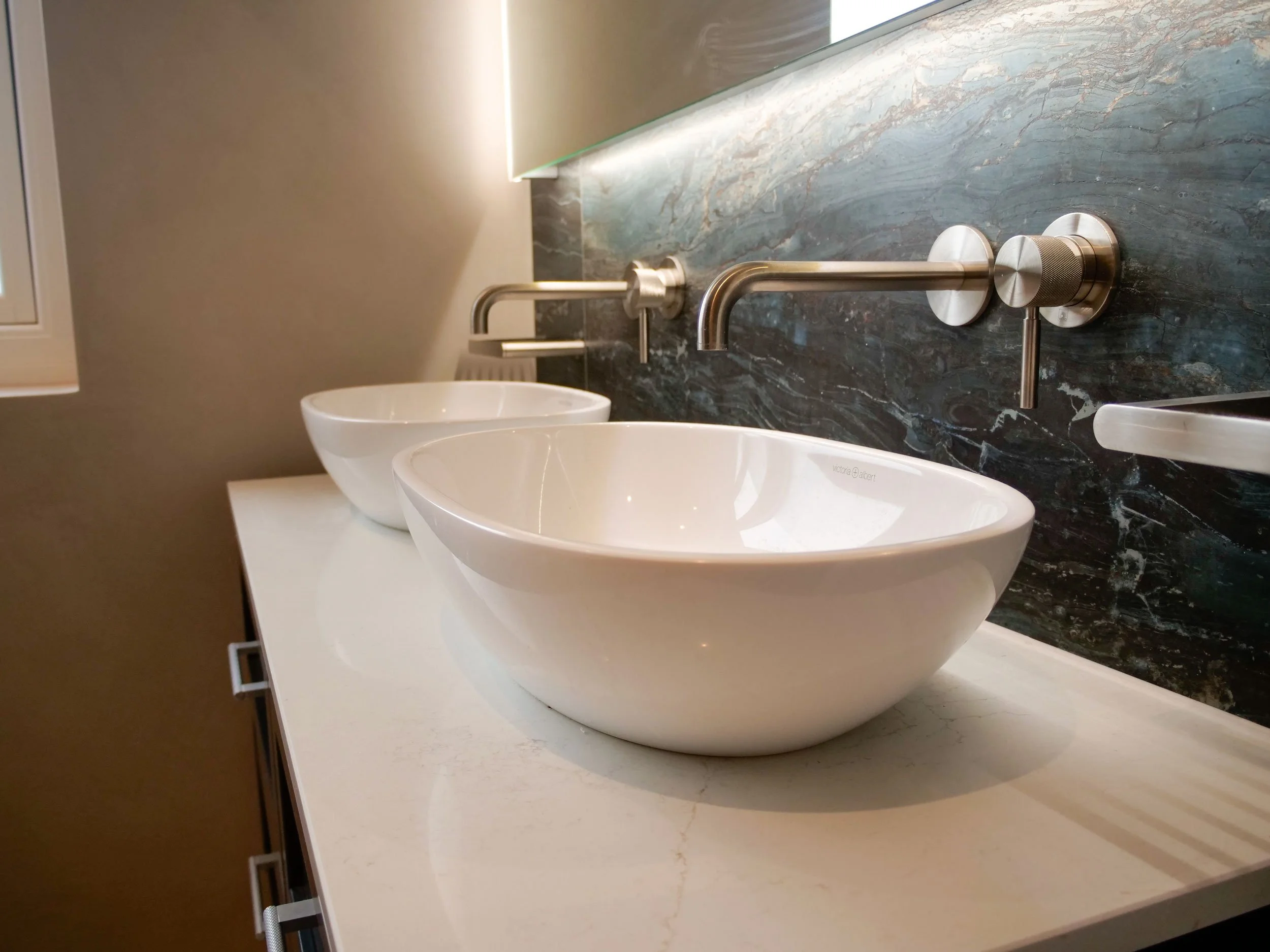Modern bathroom with two white vessel sinks mounted on a white countertop, stainless steel wall-mounted faucets, and a dark marble backsplash.