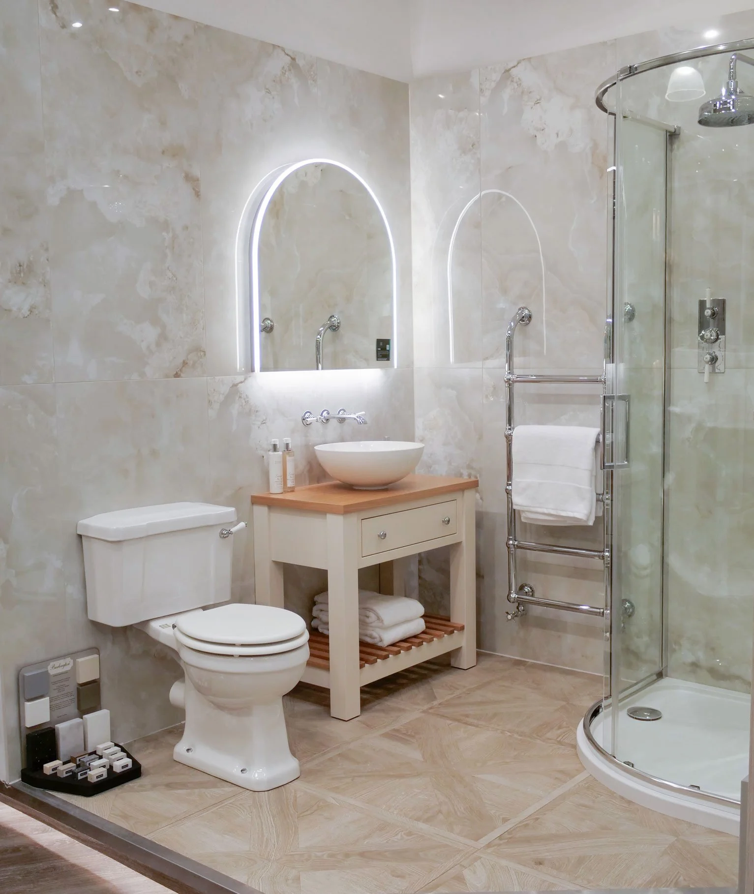 Modern bathroom with beige marble walls and floor, a white toilet, a vanity with a vessel sink, illuminated mirror, towel rack, and curved glass shower enclosure.