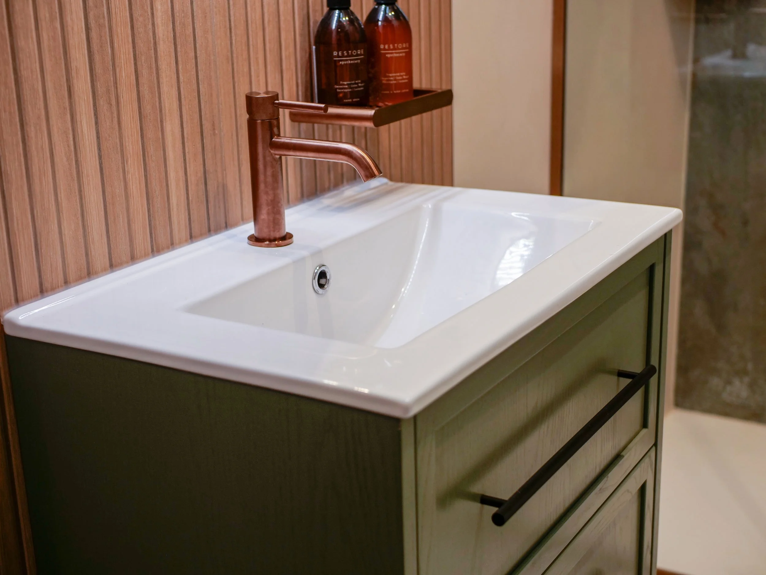 Bathroom sink with a copper faucet, a green cabinet, and a wooden wall behind, with two brown bottles on a shelf above.