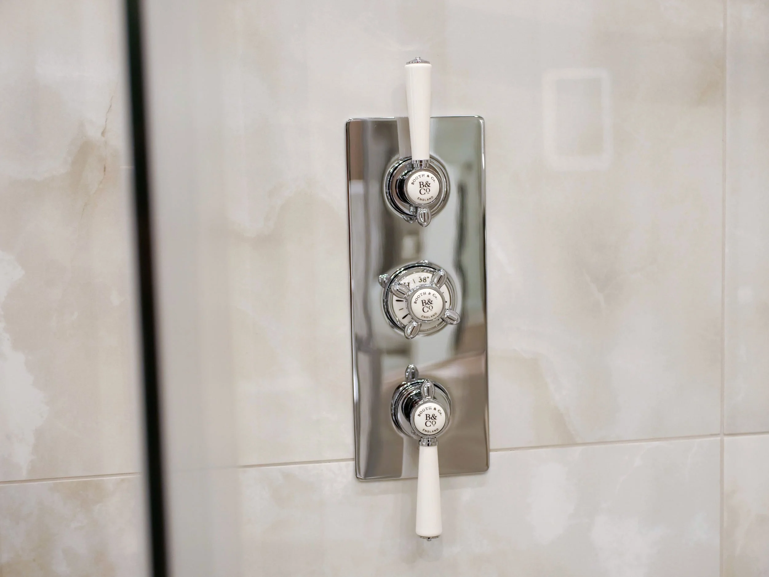Close-up of a shower control panel with three metallic knobs and white handles, mounted on a tiled bathroom wall.