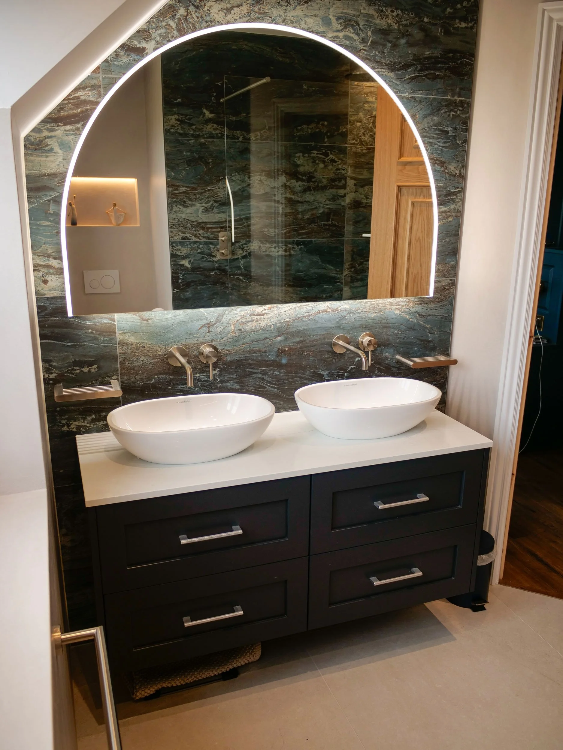 Modern bathroom with dual vessel sinks on a white vanity, large backlit arched mirror, dark marble wall behind the mirror, mirrored shower with glass door, and wall-mounted faucets.
