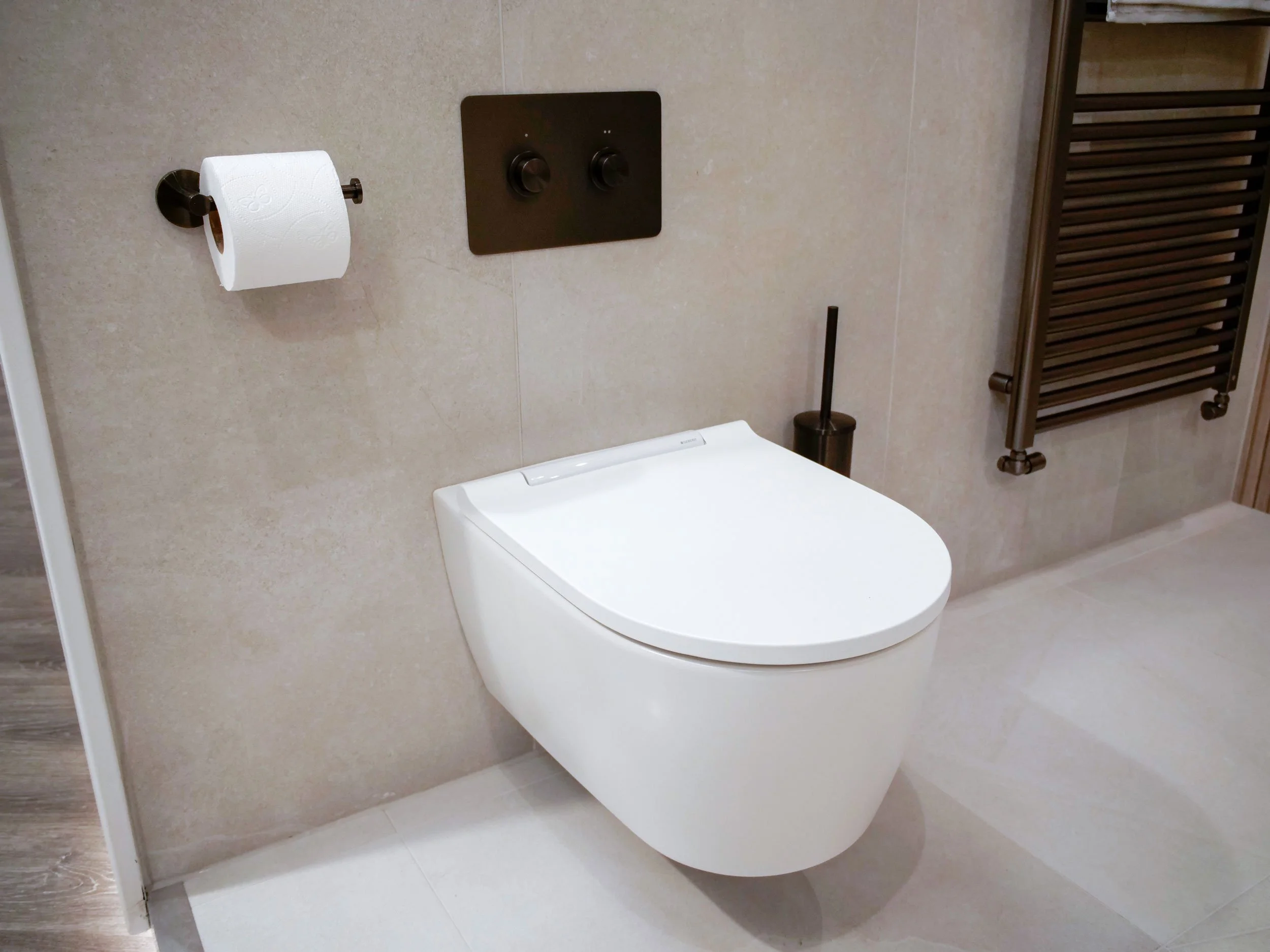 Modern bathroom with wall-mounted toilet, black toilet brush, toilet paper on a holder, black flush buttons, and a heated towel rack.