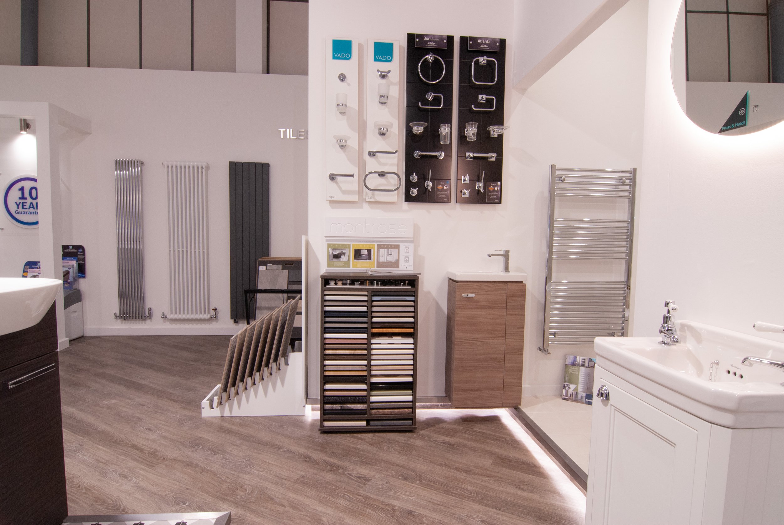 Display of bathroom fixtures and fittings in a showroom, including towel radiators, cabinet with tiles, sample tiles, a small wall sink, and a larger porcelain sink with a mirror.