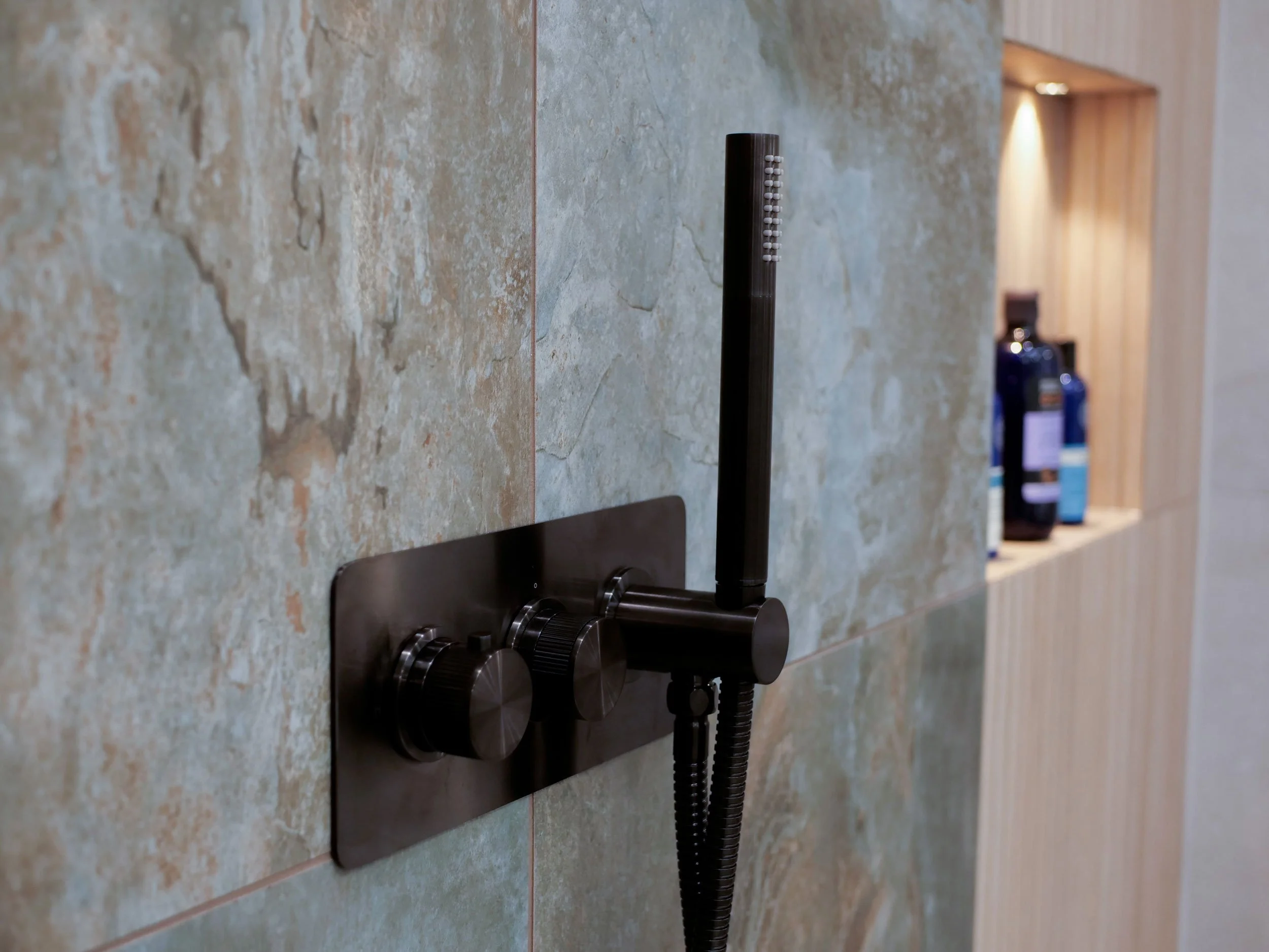 Close-up of a shower faucet with two black control knobs and a black handheld showerhead mounted on a tiled bathroom wall.