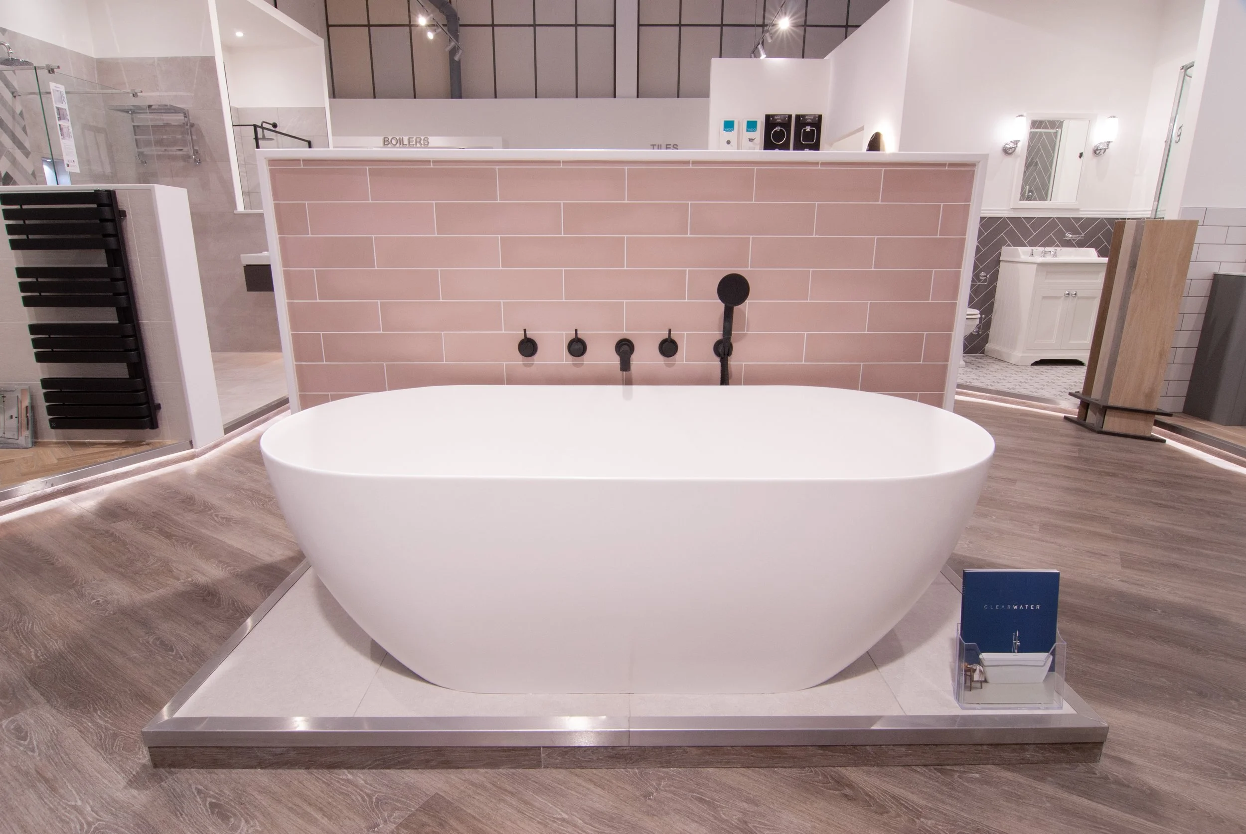 A modern bathroom with a standalone white bathtub on a raised platform, surrounded by a wood grain floor. Behind the tub is a pink-tiled wall with black fixtures, including a handheld showerhead. To the right, there is a white bathroom sink with a mi