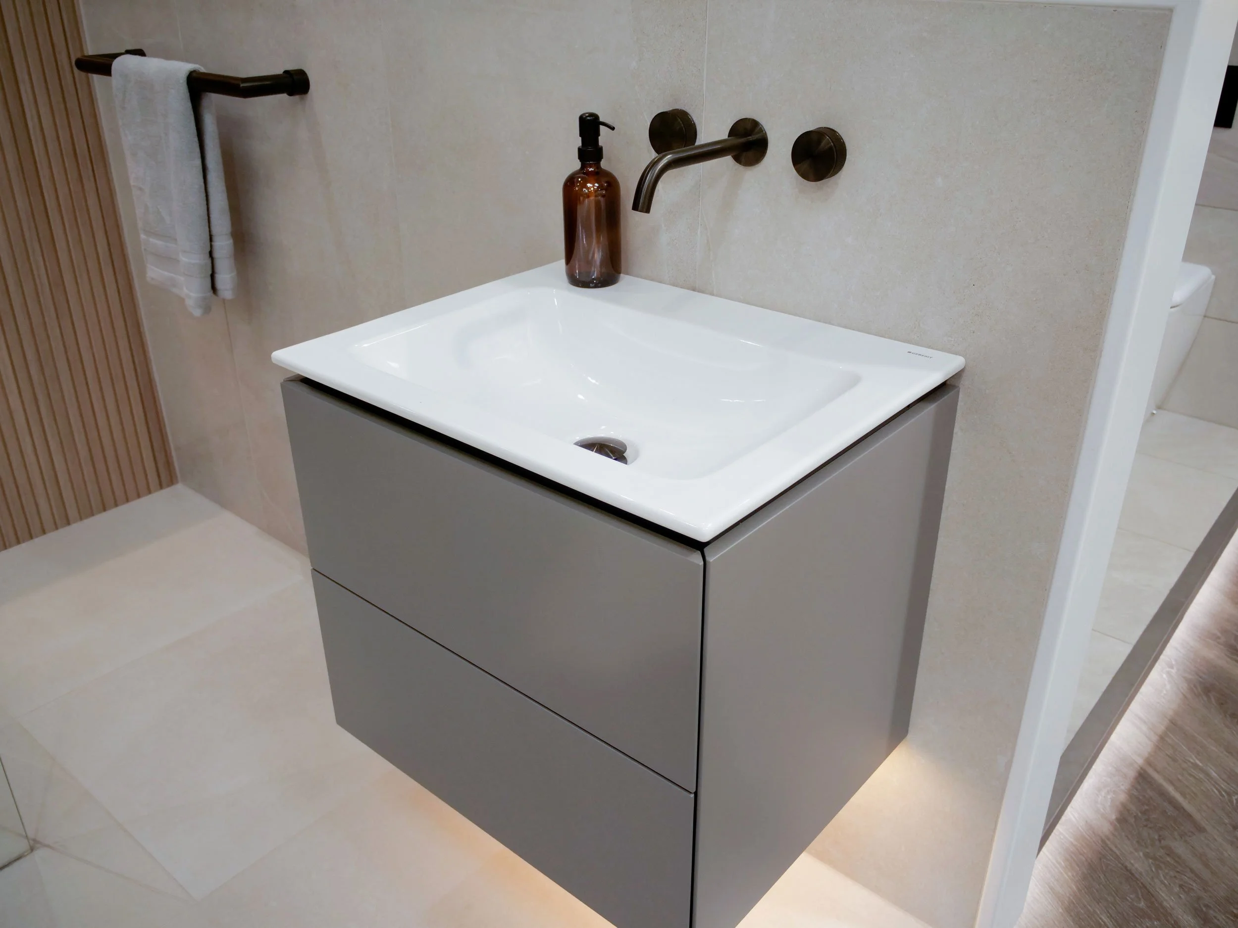 Modern bathroom vanity with a rectangular white sink, a brown soap dispenser, a wall-mounted faucet, and a towel hanging on a black towel bar nearby.