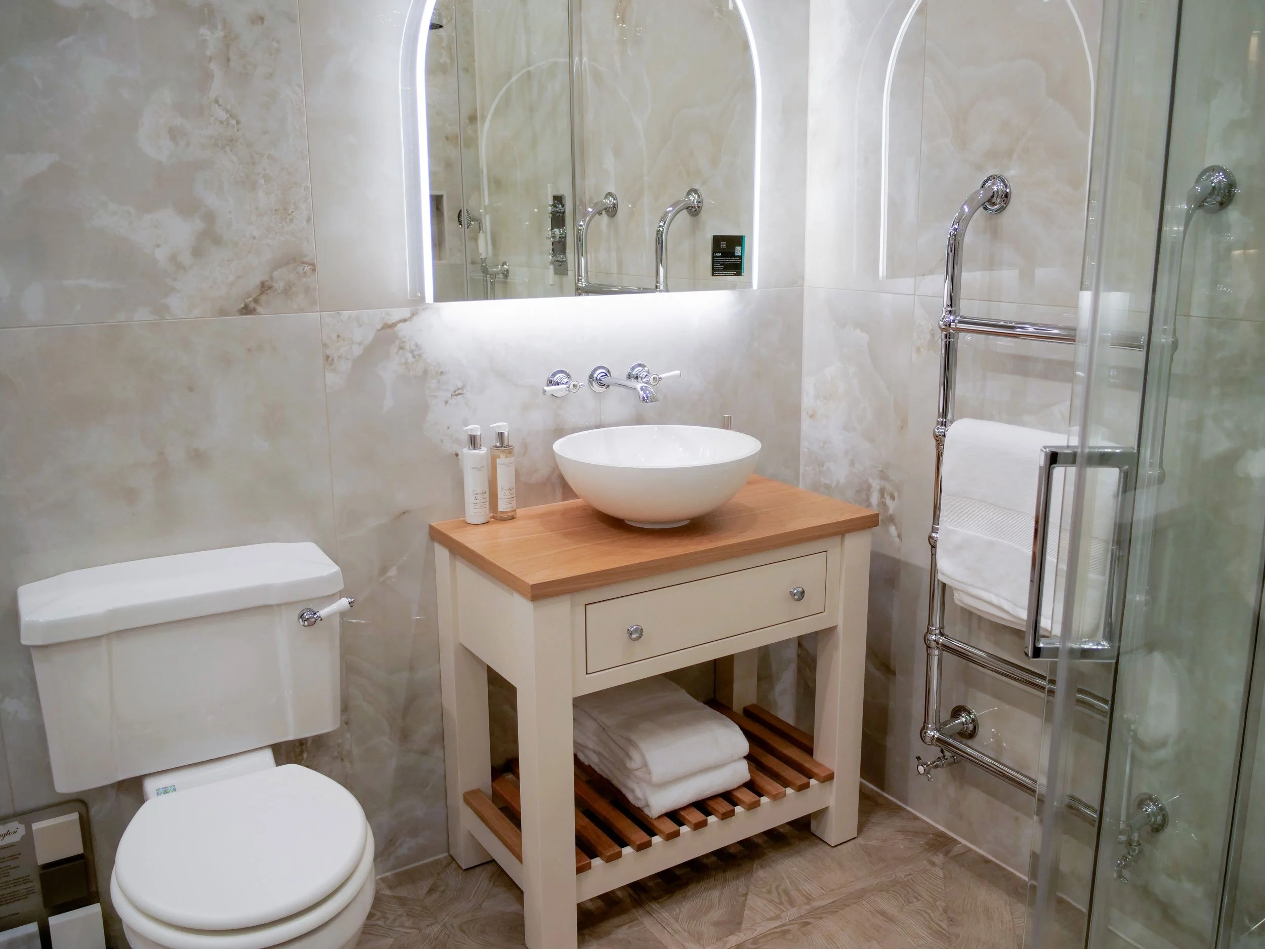 Bathroom with a beige marble wall, a white toilet, a white vanity with a wooden top and a vessel sink, a large mirror with LED lighting, and a chrome towel warmer with a white towel, and a glass shower enclosure.