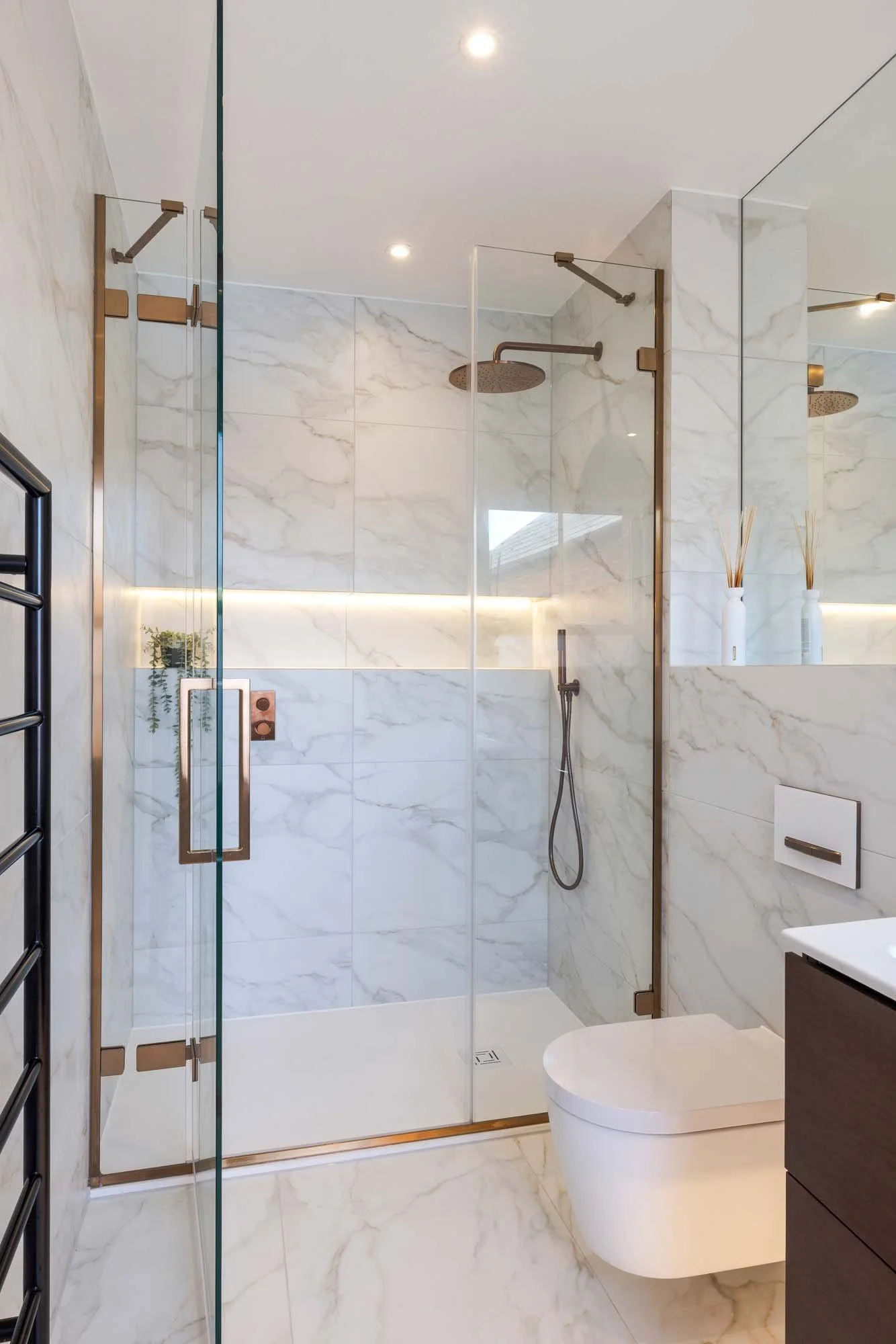 Modern bathroom with marble walls and floor, glass shower enclosure with brass fixtures, black towel warmer on the left, and a white wall-mounted toilet next to a dark vanity.