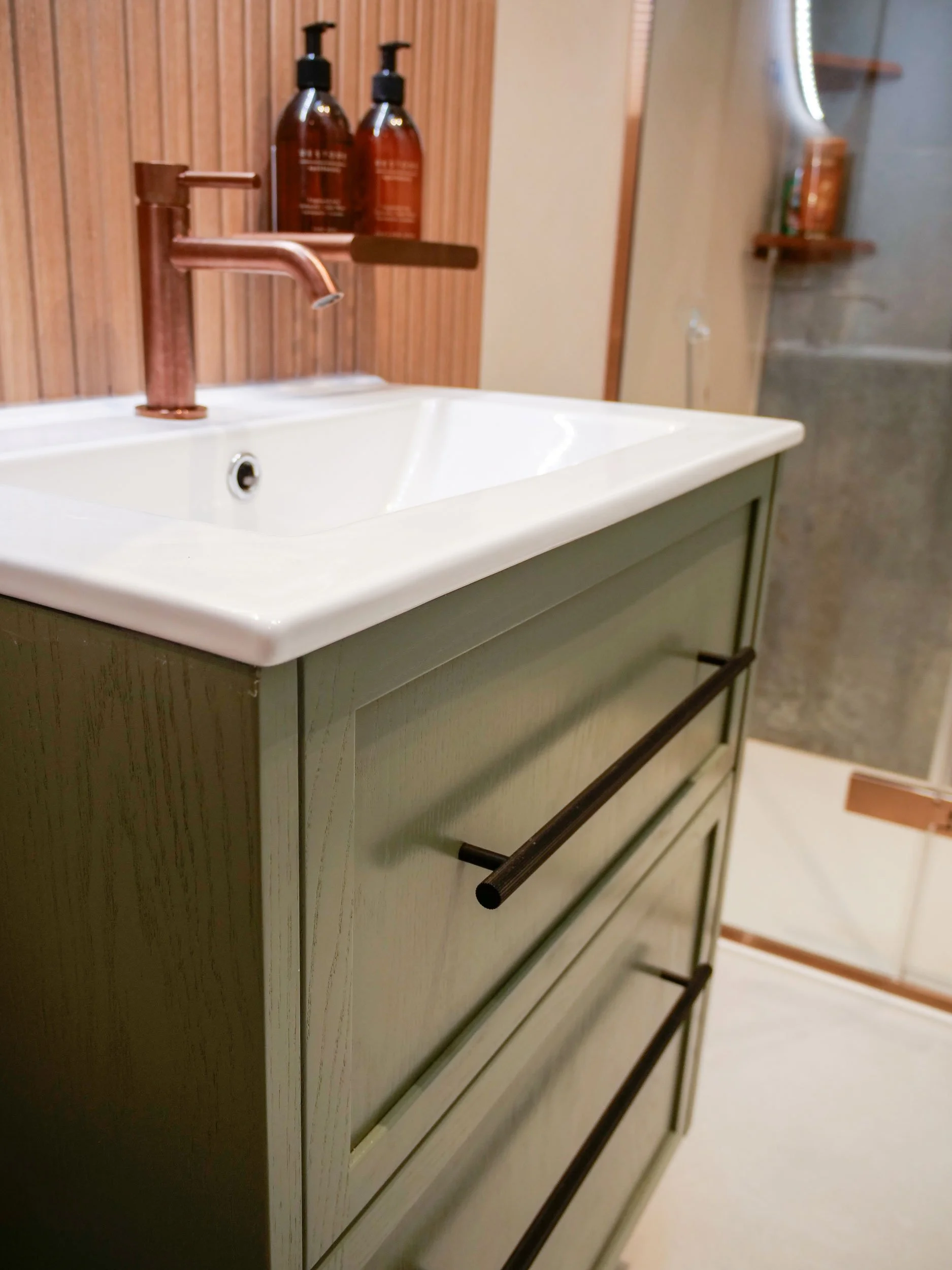 Close-up of a bathroom sink vanity with a copper-colored faucet and drawers. Behind the sink, there are two amber-colored soap or lotion bottles on a wooden shelf.