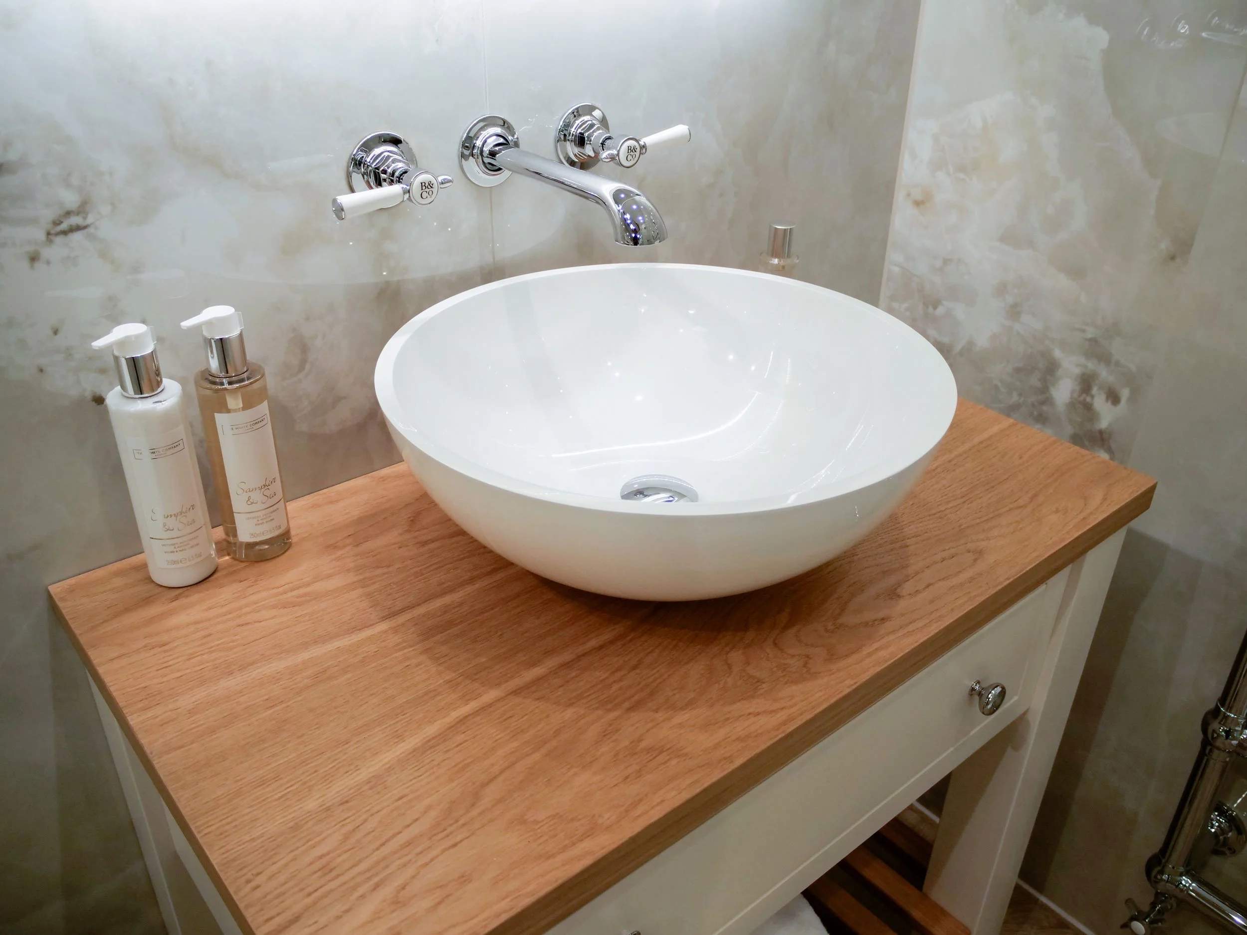 Bathroom sink with a white round vessel basin, a chrome faucet, and soap bottles on a wooden countertop against a marble wall.