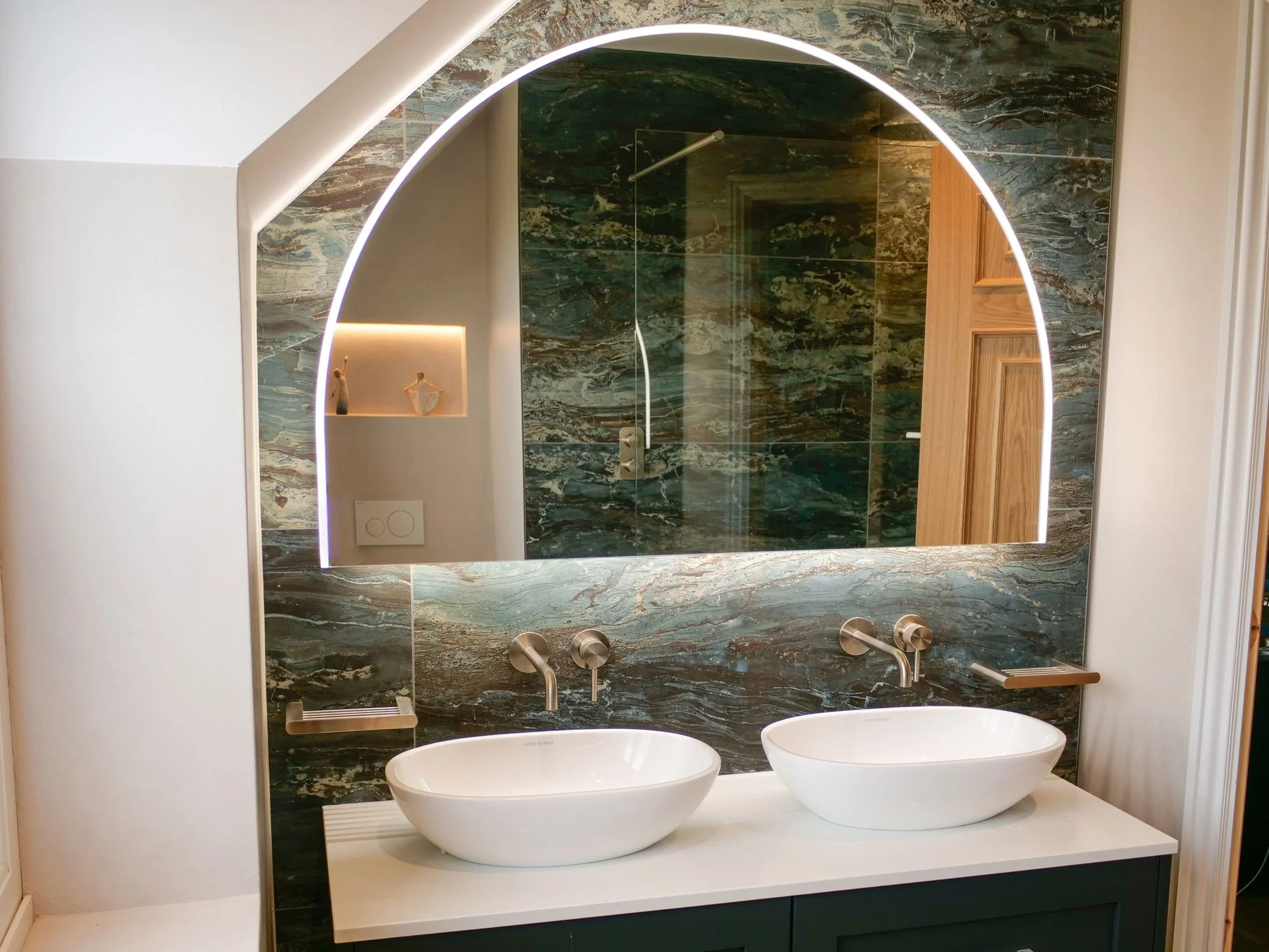 Modern bathroom vanity with two oval vessel sinks, wall-mounted faucets, a large arched mirror with backlighting, and a marble wall with green and brown veining. Reflection shows shower area and wall niche with decor.