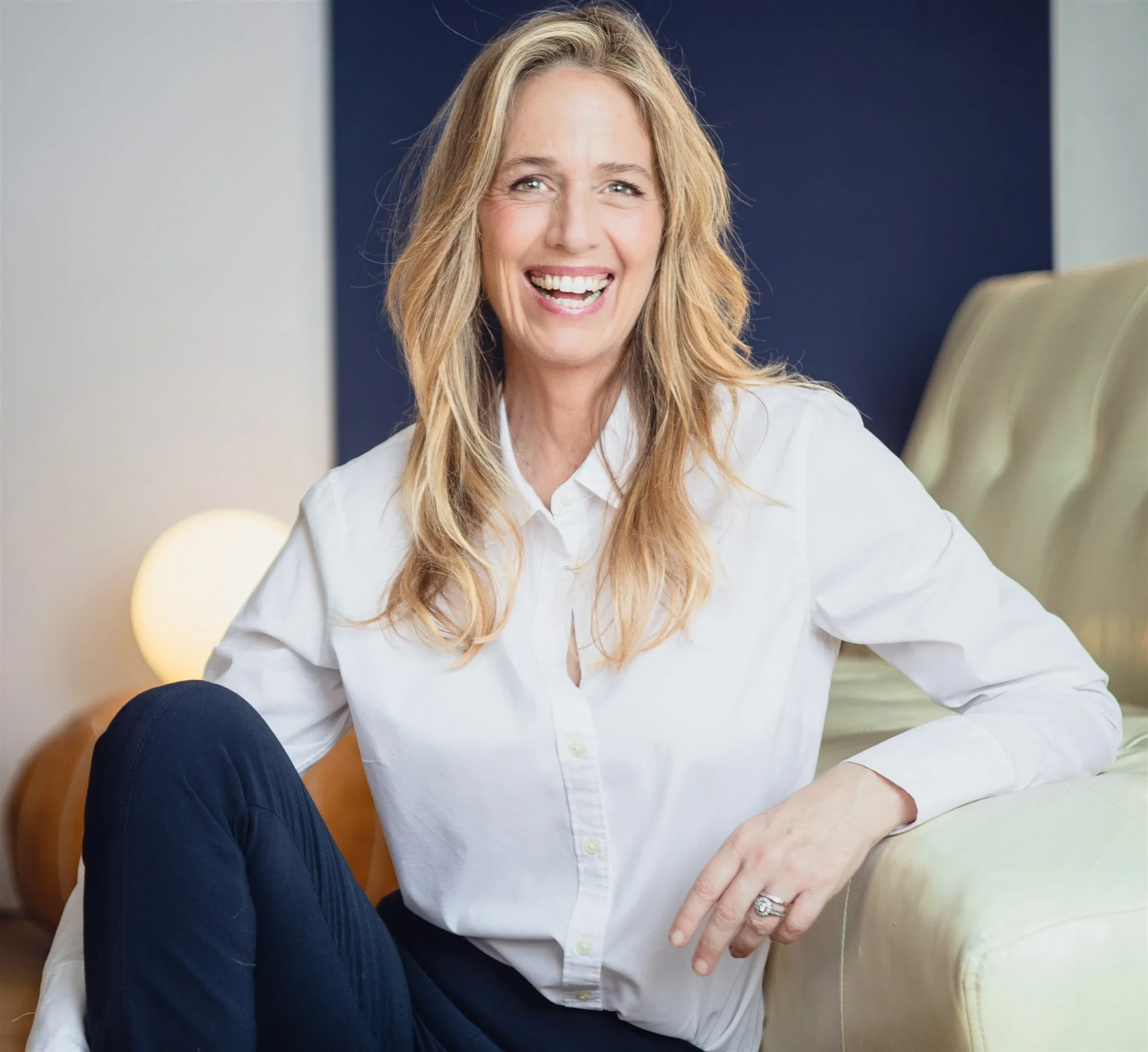 A woman with long blonde hair and a white shirt smiling while sitting on a sofa in a modern living room.