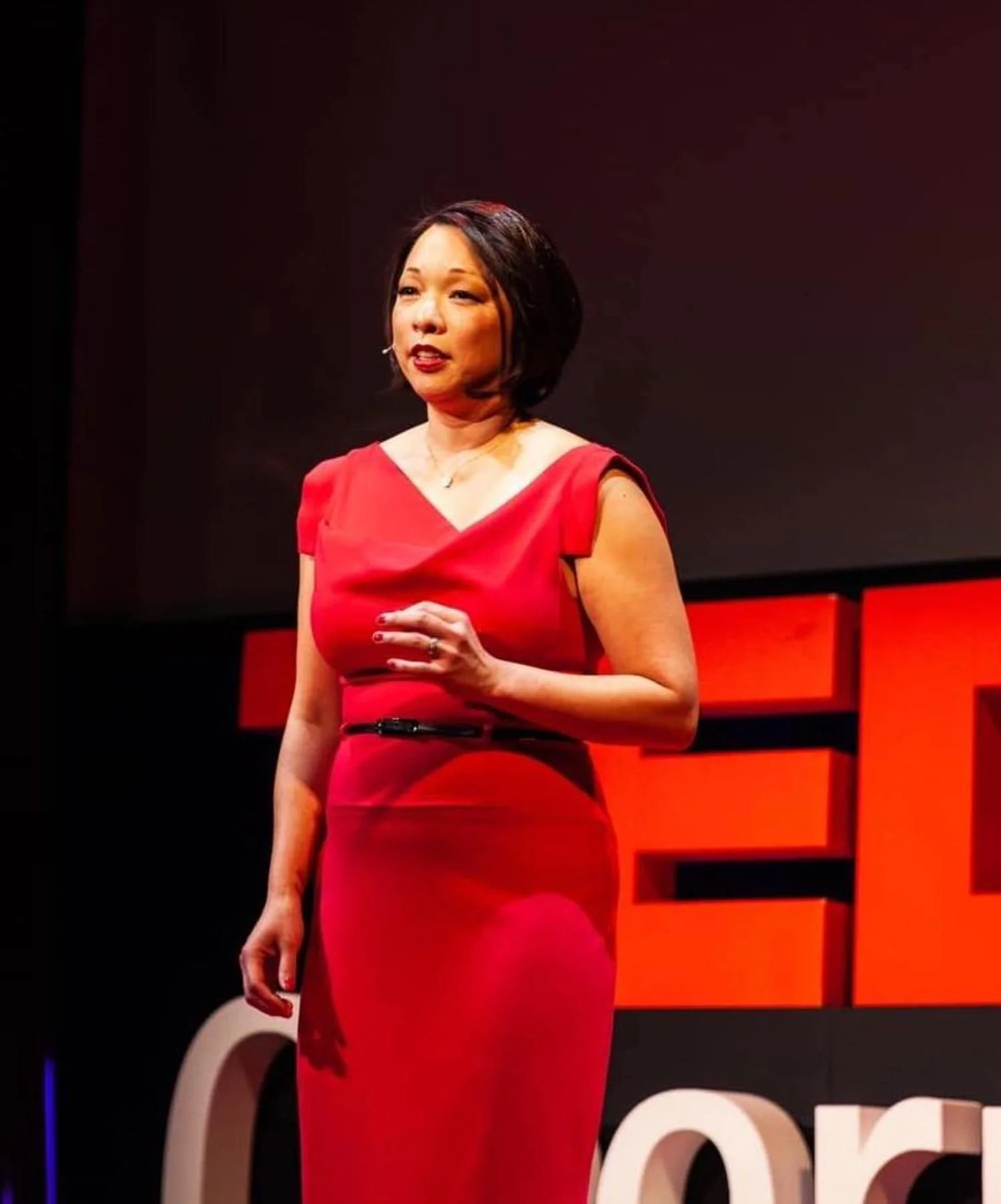 A woman in a red dress giving a talk on a stage with red and black background elements.
