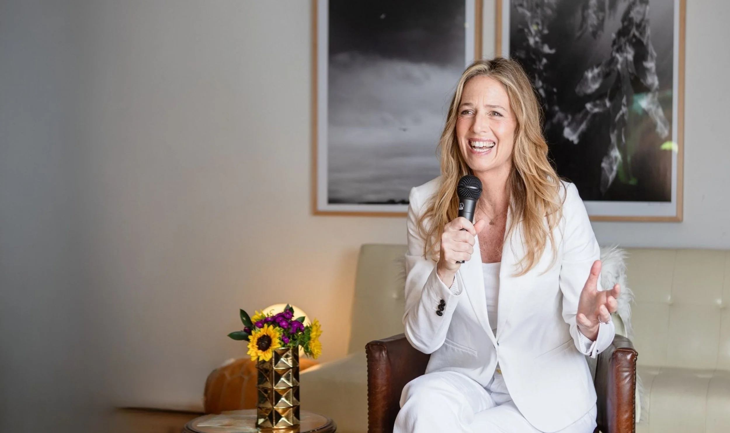 A woman in a white suit sitting on a chair, holding a microphone, and speaking or singing with a smile. There is a small table with a vase of colorful flowers beside her, and framed artwork hanging on the wall behind her.