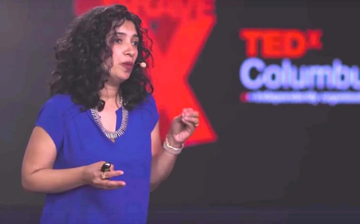 A woman with curly dark hair wearing a blue dress and a silver necklace giving a presentation on a stage with a TedX Columbia backdrop.
