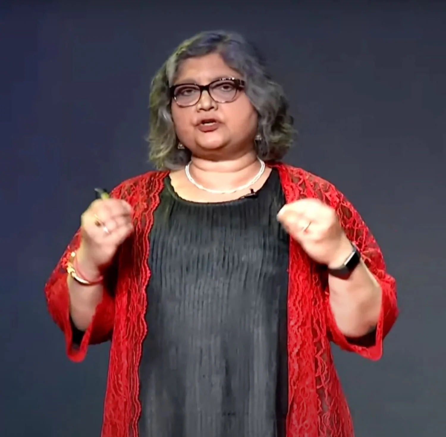 Woman with gray hair, glasses, wearing a black top and a red lace jacket, speaking during a presentation or speech.