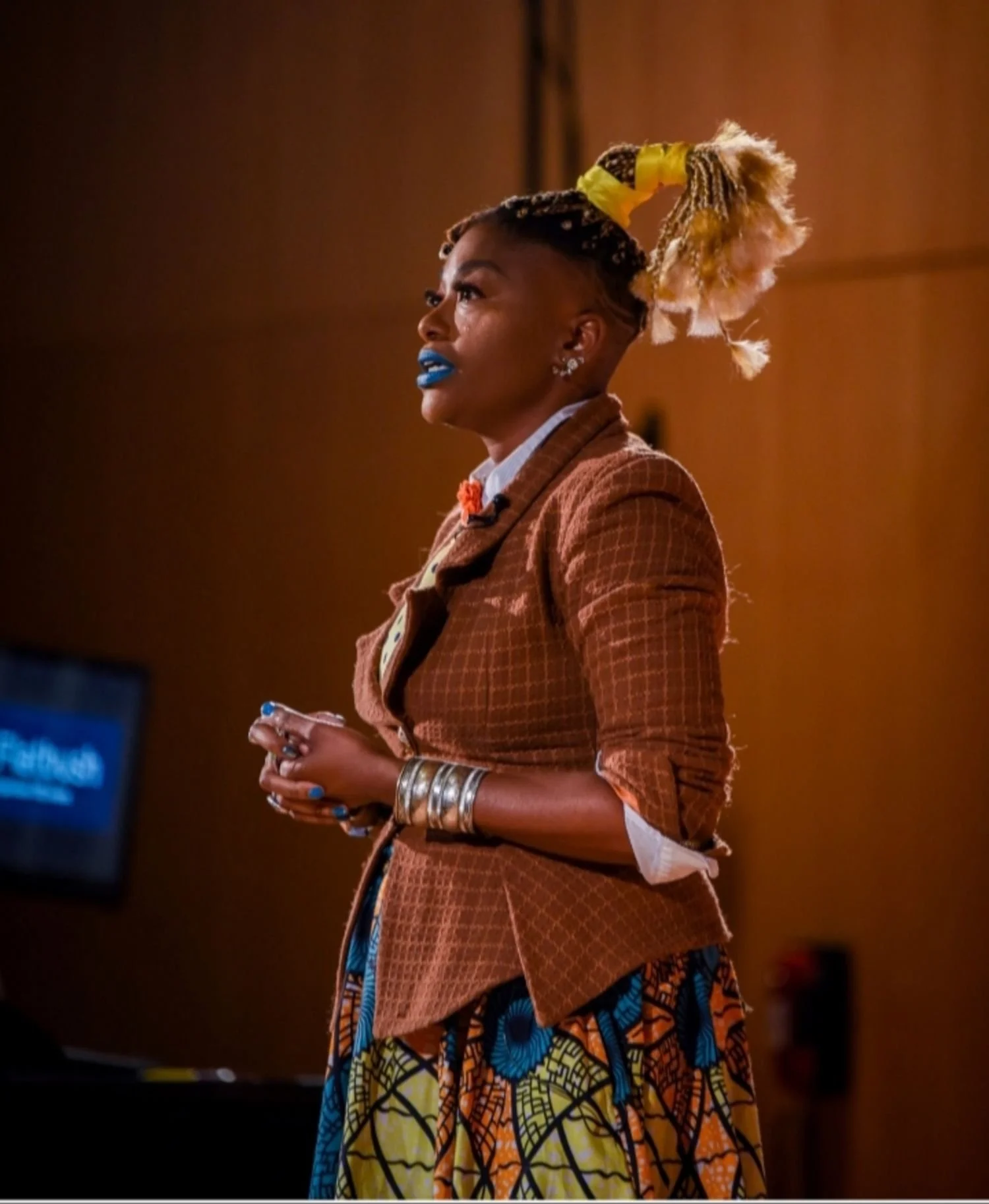 A woman with colorful braided hair tied in a high ponytail, wearing a brown checkered blazer, multicolored African print skirt, and silver bracelets, standing in profile against a brown background.