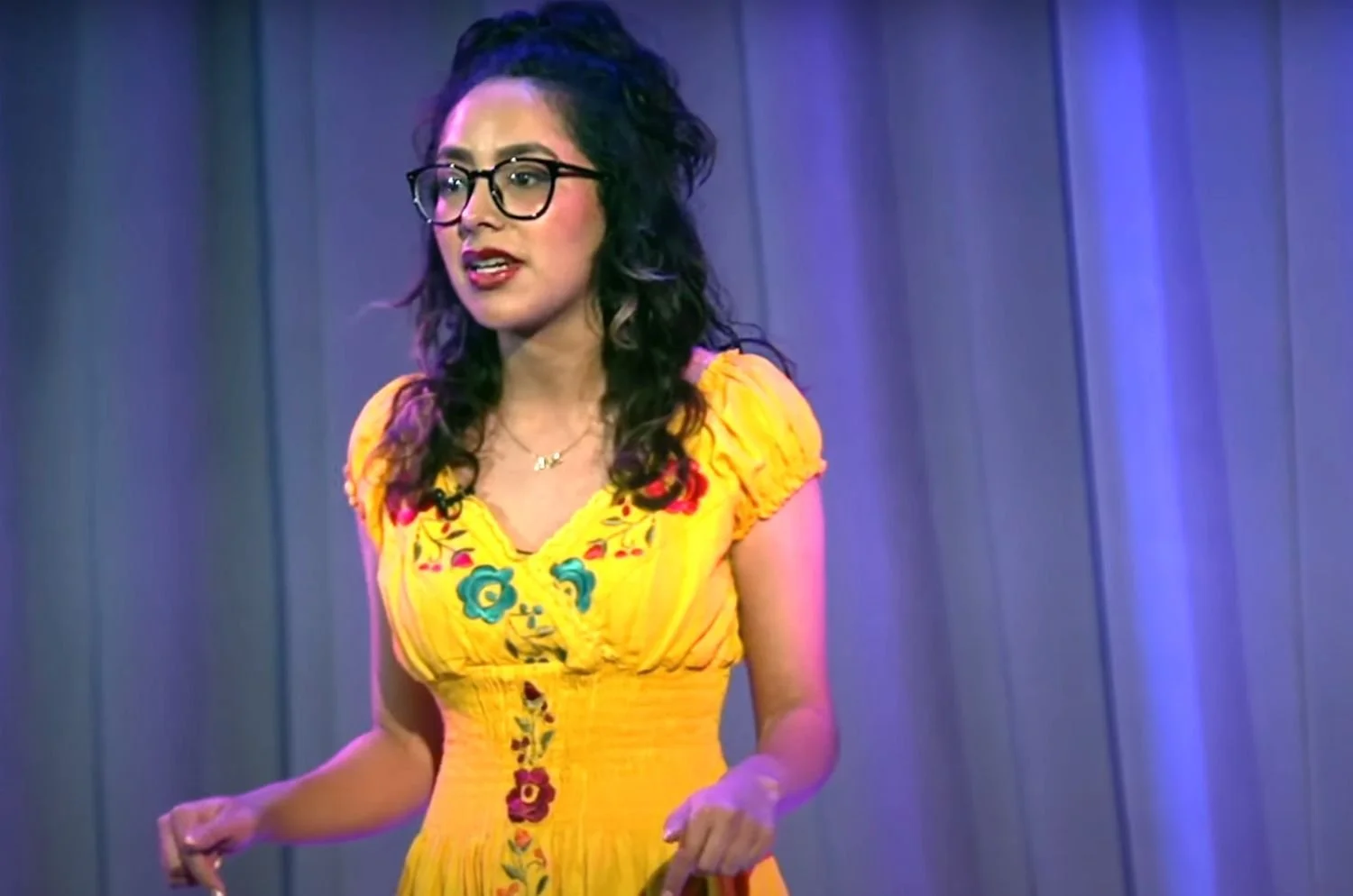 A woman with dark curly hair, glasses, and wearing a yellow embroidered dress stands in front of a curtain backdrop.