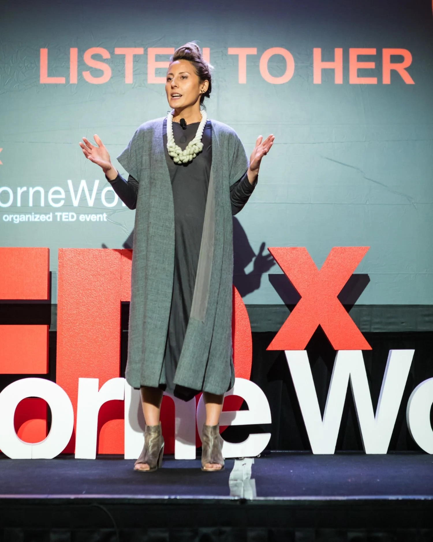 A woman speaking on stage at a TEDx event, with large red and white TEDx letters behind her and a screen that reads 'Listen to Her'.