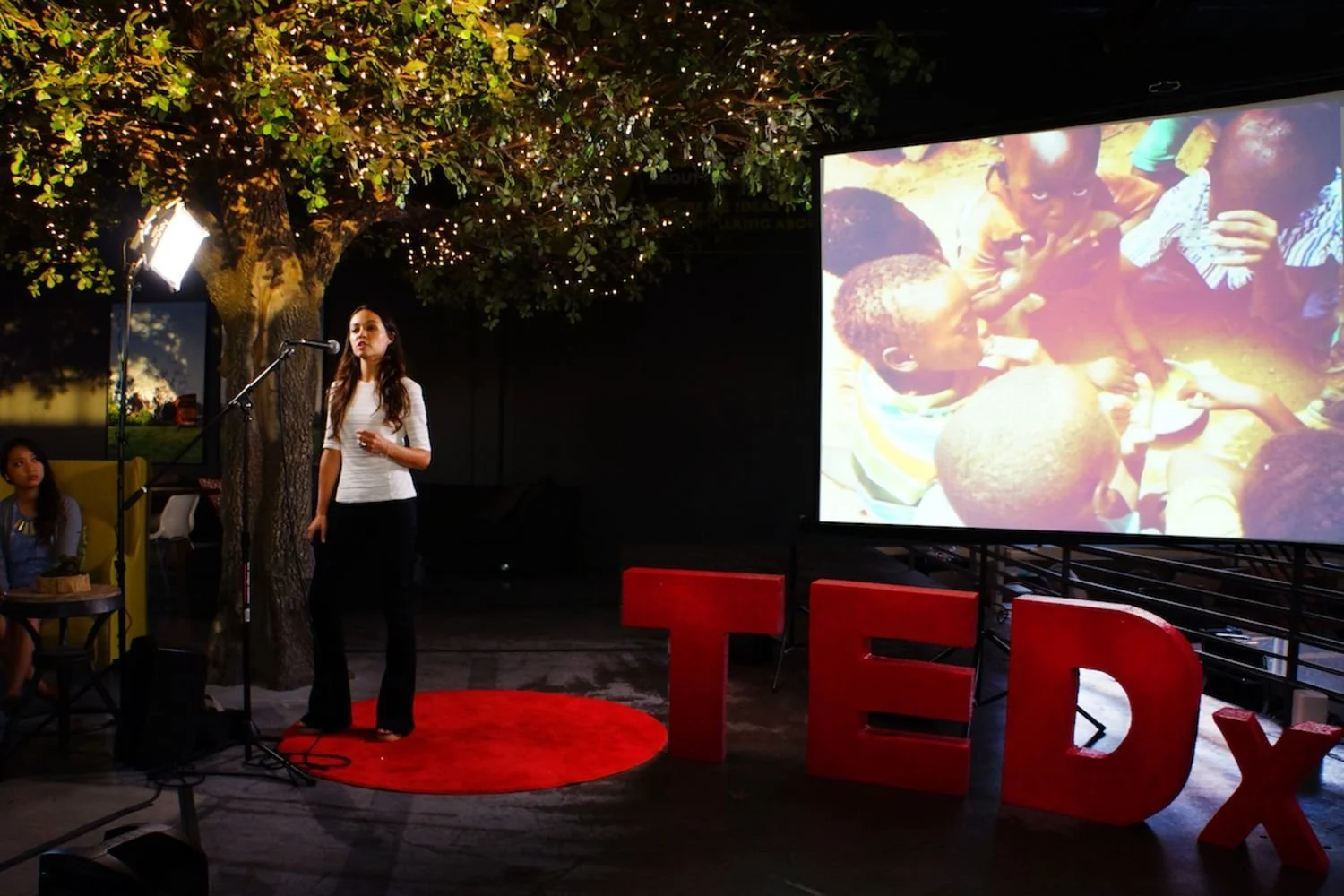 A woman giving a presentation at a TED event with a large screen displaying a photo of children in a rural setting. There is a red TED sign and a woman sitting nearby under a lit tree.