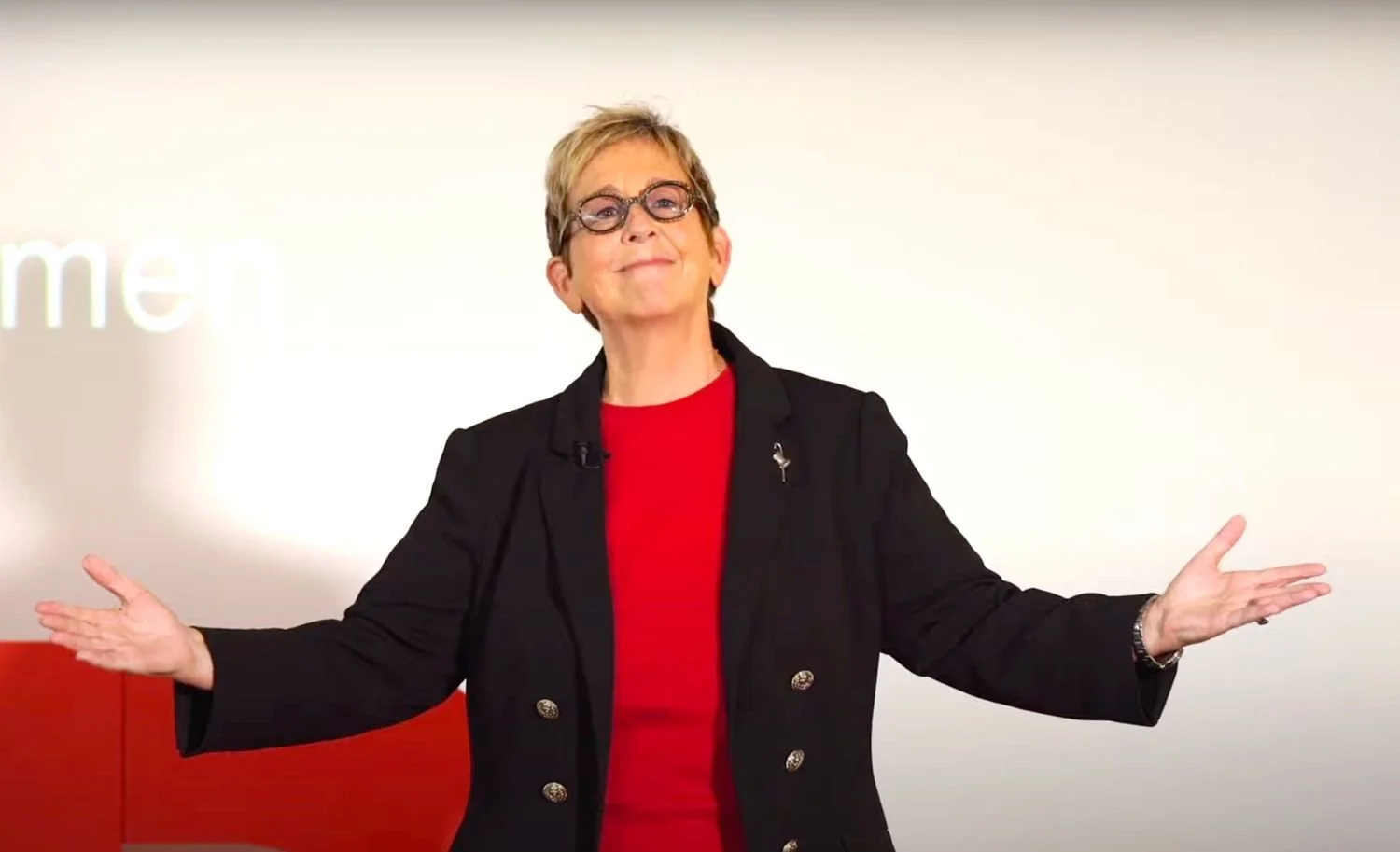 A woman with short blonde hair, glasses, wearing a red top and black jacket, standing with arms open in front of a white background.