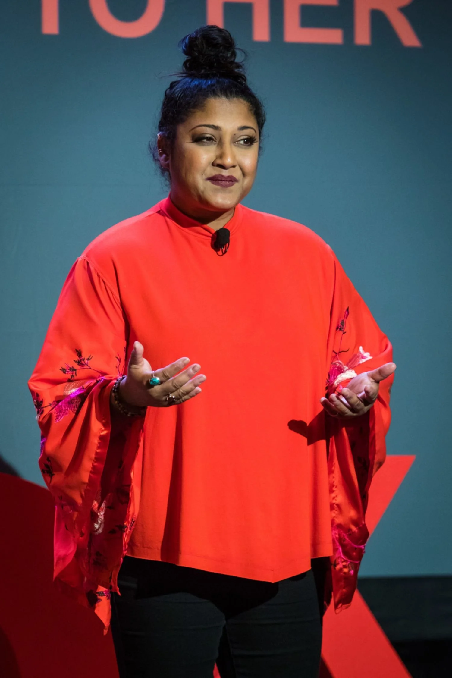 A woman with dark hair tied in a bun, wearing a red top with floral embroidery on the sleeves, standing on a stage with a blue background that has red text. She's gesturing with her hands and has a small microphone attached to her shirt.