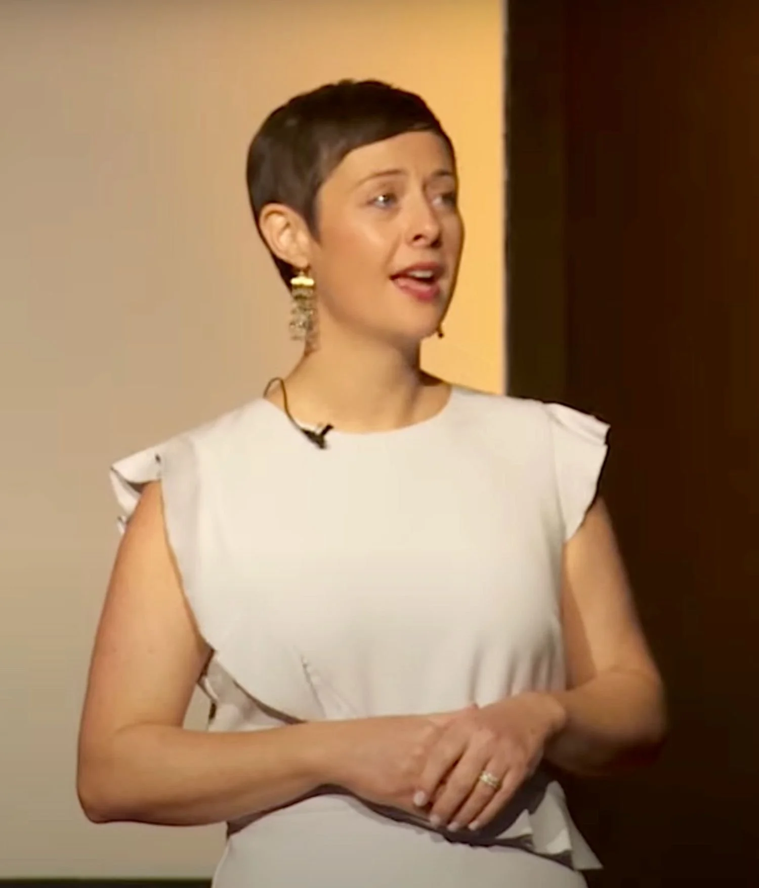 A woman with short dark hair wearing a white sleeveless top, gold earrings, and a ring, standing and speaking in front of a beige and dark brown background.