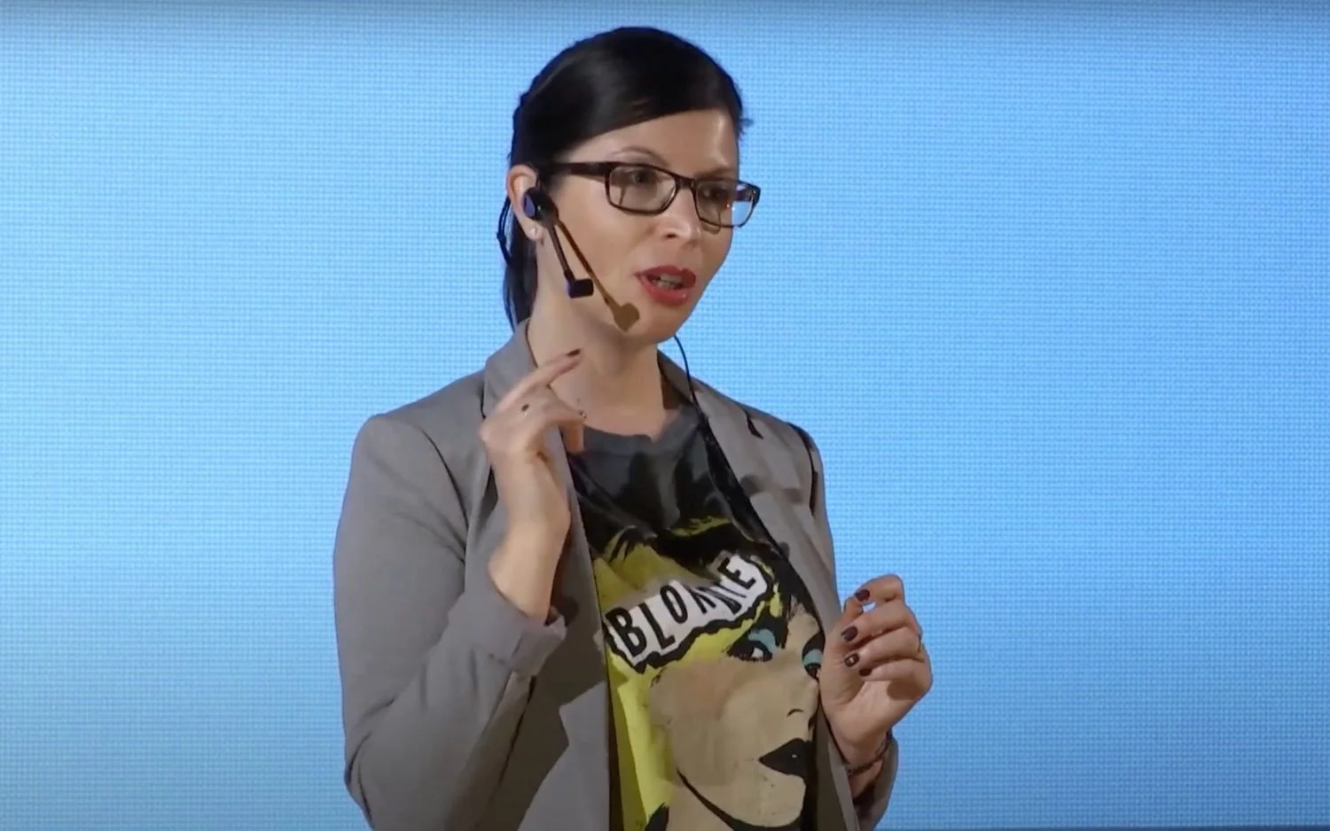 A woman wearing glasses and a gray blazer speaking into a headset microphone during a presentation, against a large blue screen background. She is wearing a graphic t-shirt with an illustrated woman's face and the word "BLOOM".