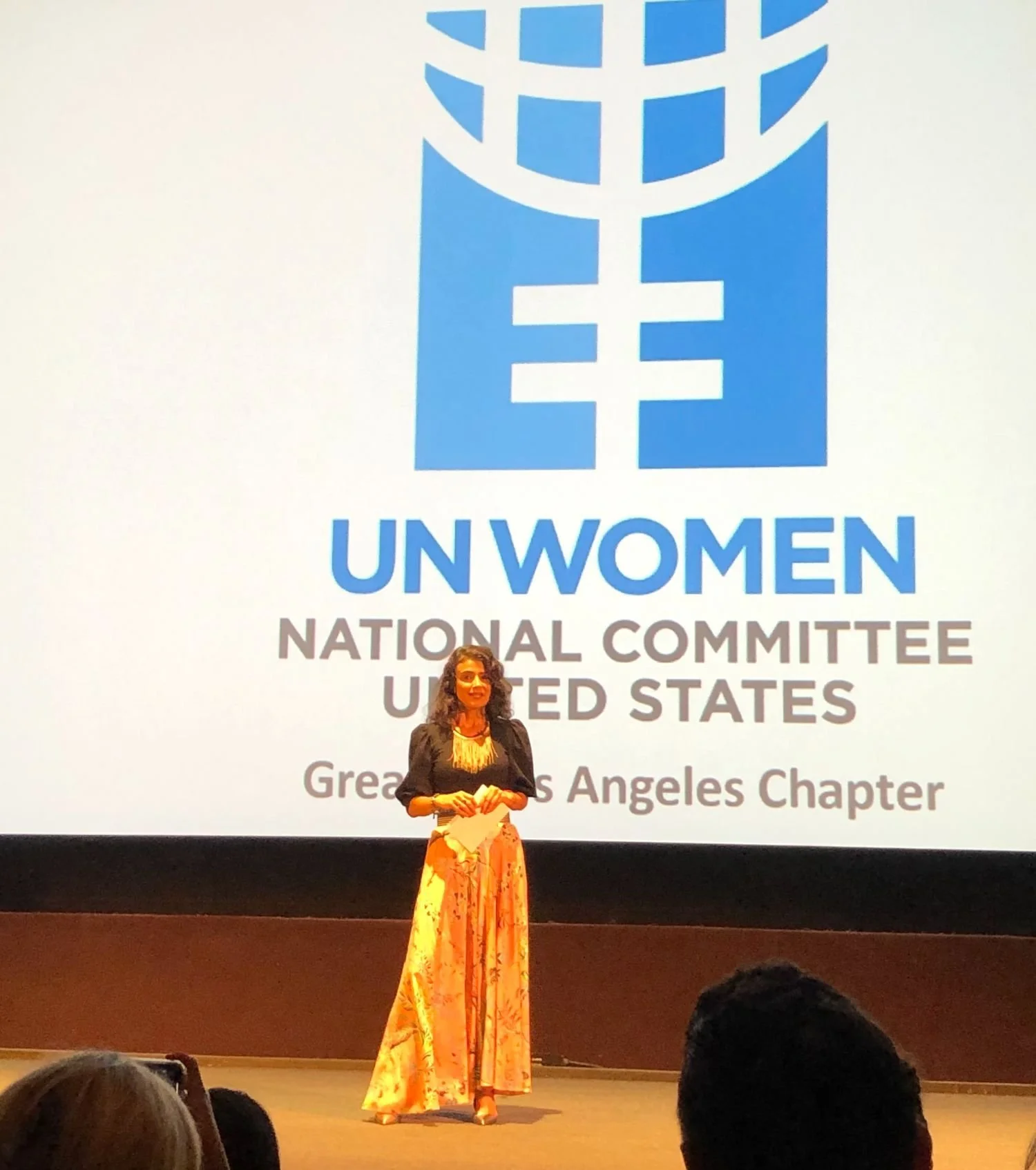 A woman standing on stage in front of a large screen displaying the UN Women logo and text, including "UN Women, National Committee, United States, Greater Los Angeles Chapter." She is holding papers and dressed in a black top with a long, colorful s
