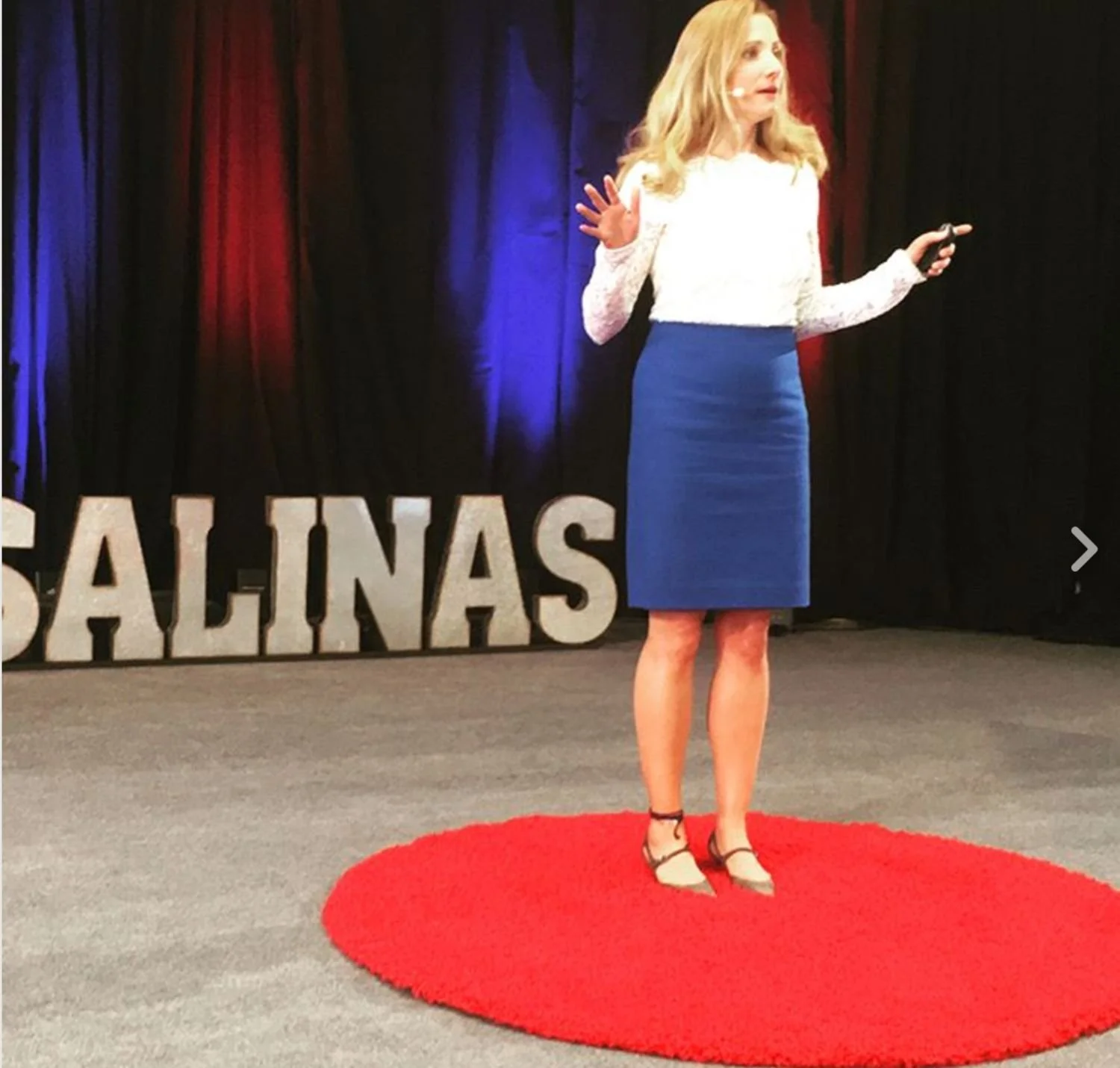A woman giving a presentation on stage, standing on a red circular carpet, with "SALINAS" spelled out in large block letters behind her, and a screen with a TEDx logo visible.