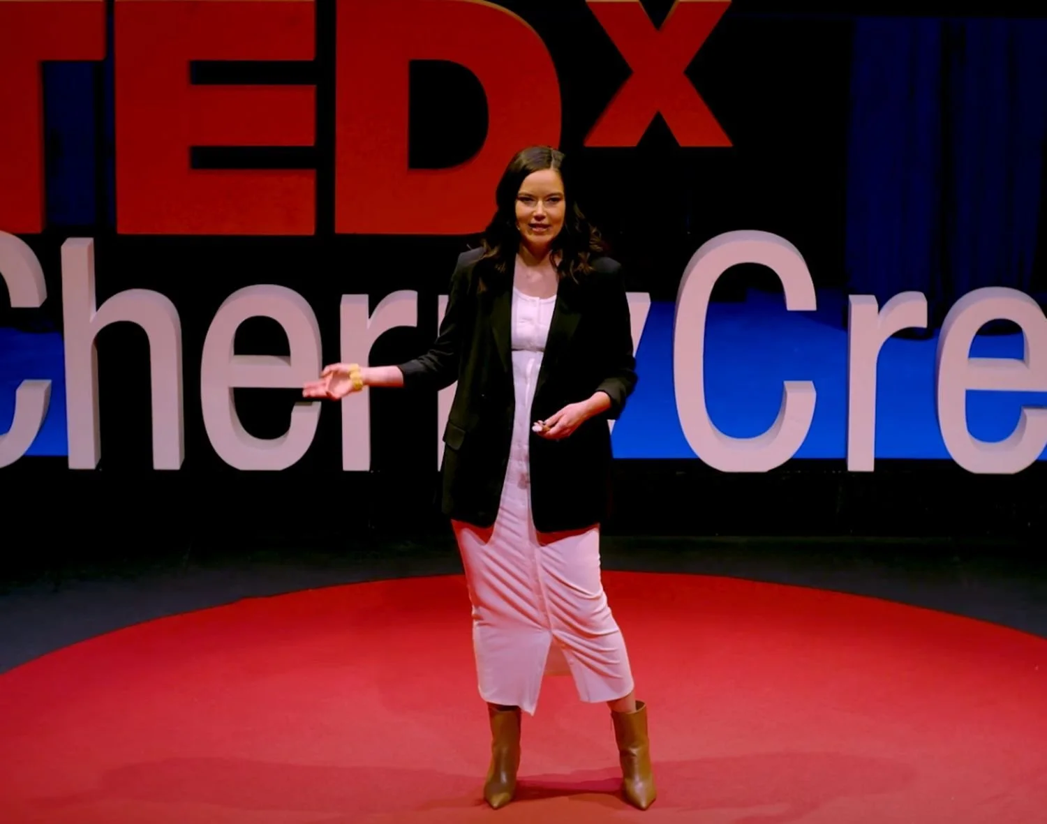 Woman giving a speech on stage with a large red and black TEDx sign and large white letters spelling 'Cherie Cre' behind her, standing on a red circular platform.