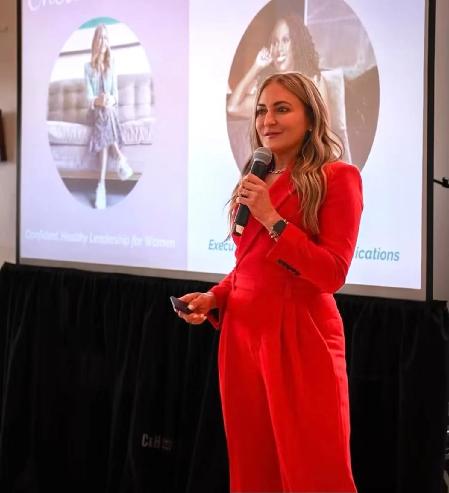 A woman in a red suit speaking into a microphone during a presentation, with a screen behind her displaying three photos of women and text related to leadership for women.