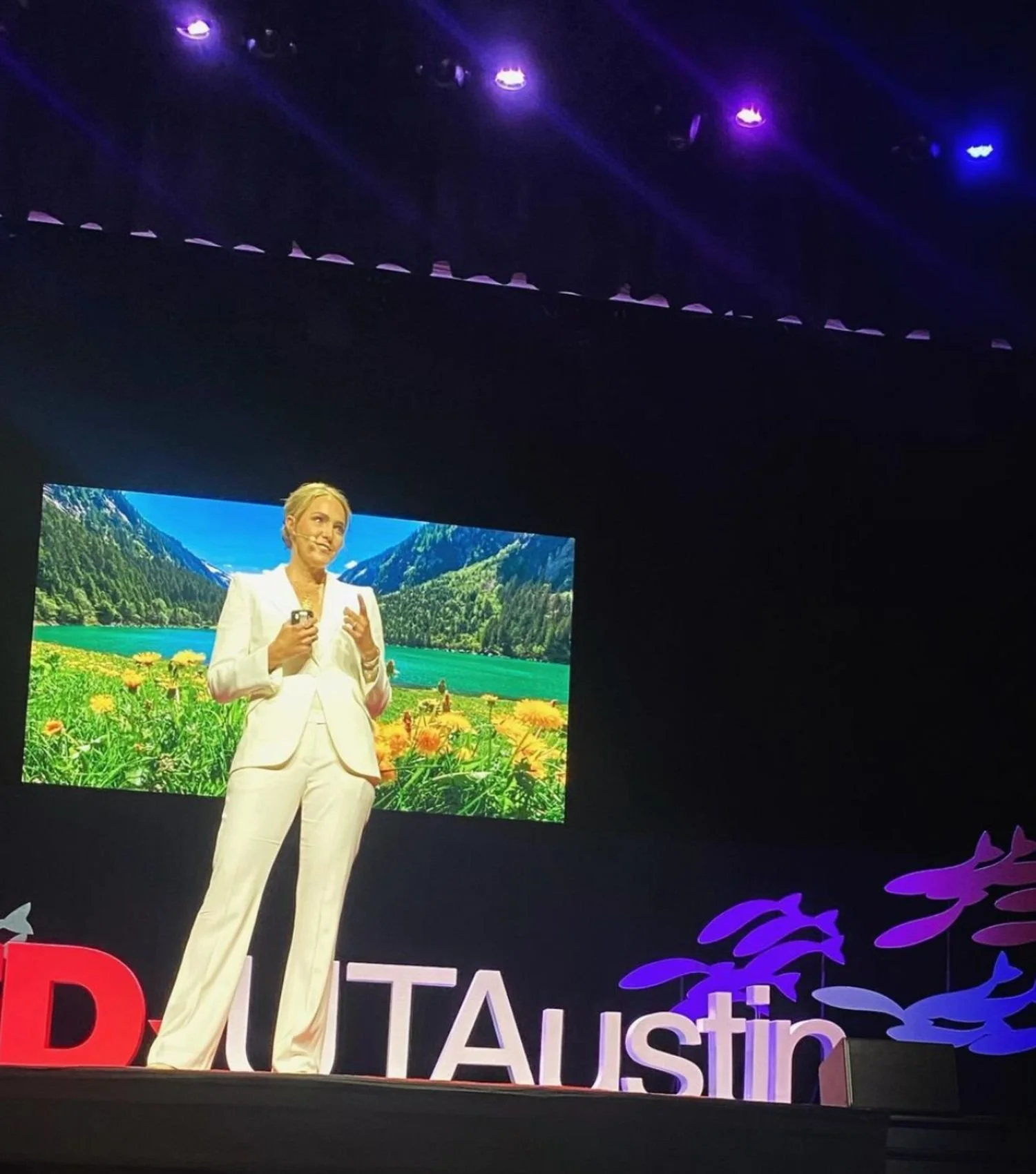 A woman giving a presentation on stage with a large screen behind her showing a scenic mountain and lake landscape. The stage has colorful lights and the partial words 'TEDxUTAustin' in large letters.
