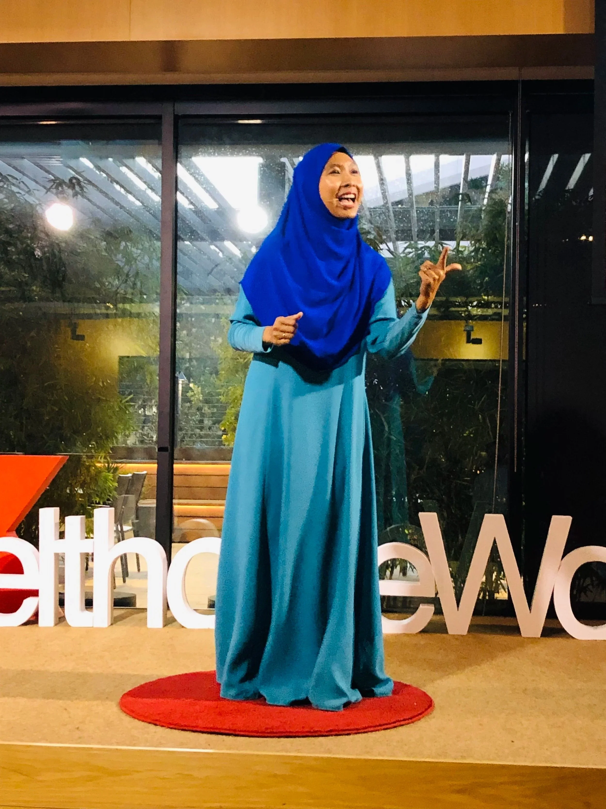 Woman in a long blue dress and blue hijab standing on a red carpet, smiling and gesturing, in front of a glass window with plants behind it.
