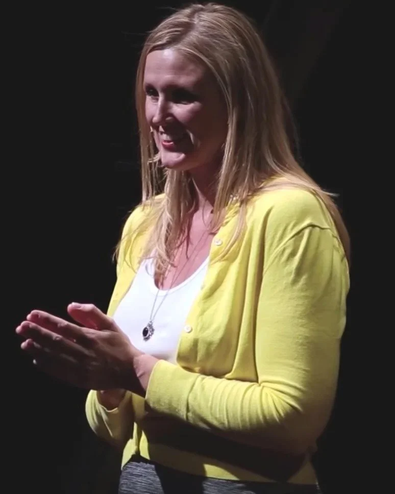 A woman with long blonde hair wearing a yellow cardigan over a white shirt, smiling and clasping her hands, standing against a dark background.