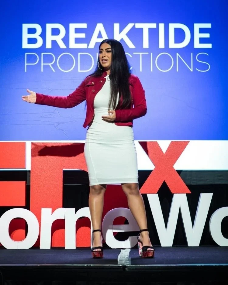 A woman standing on stage in front of a TEDx logo, wearing a white dress, red jacket, and high heels, gesturing with one arm extended.