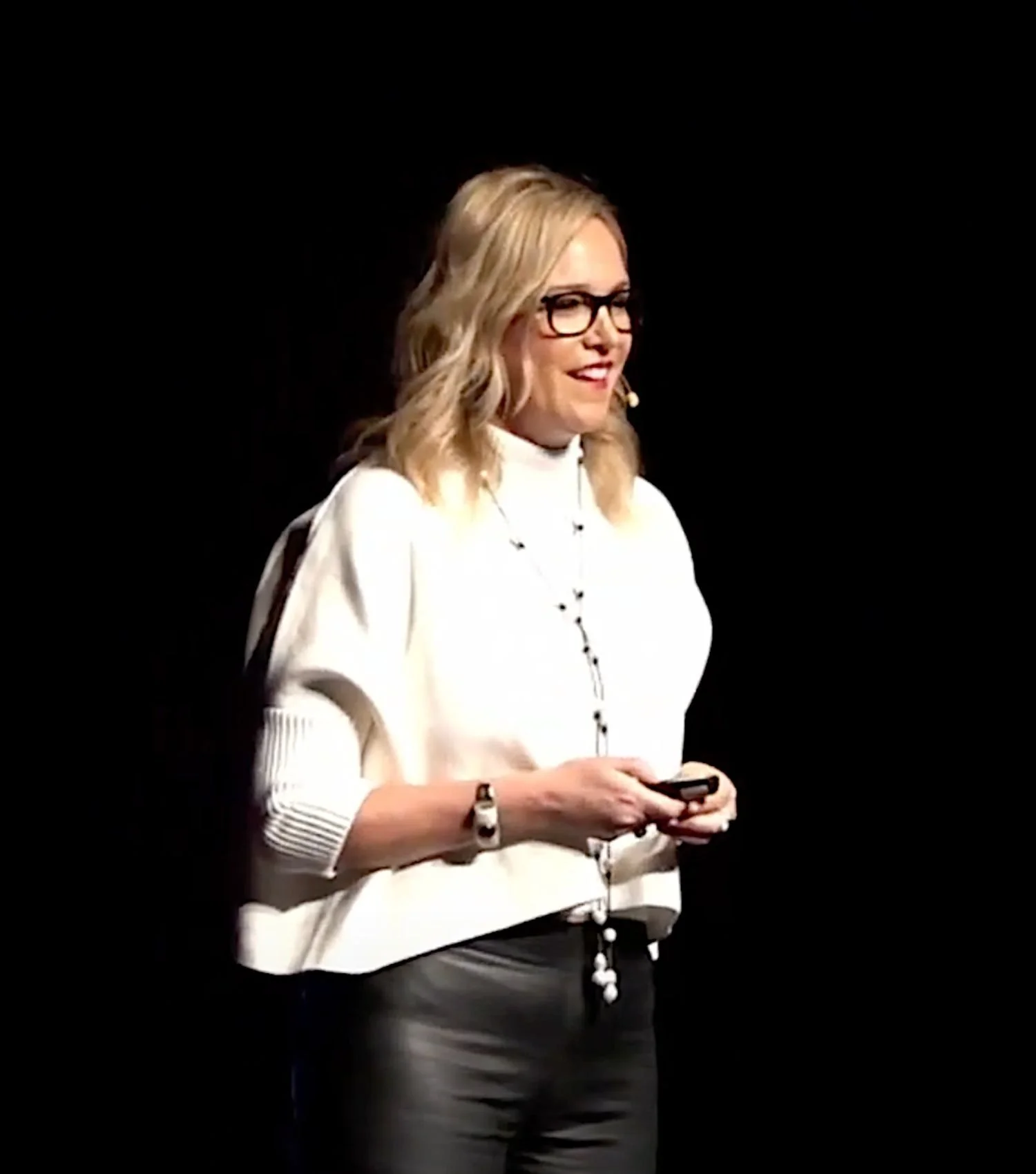 A woman with blonde hair, black glasses, and wearing a white sweater and black pants, standing on stage with a dark background, holding a small device.