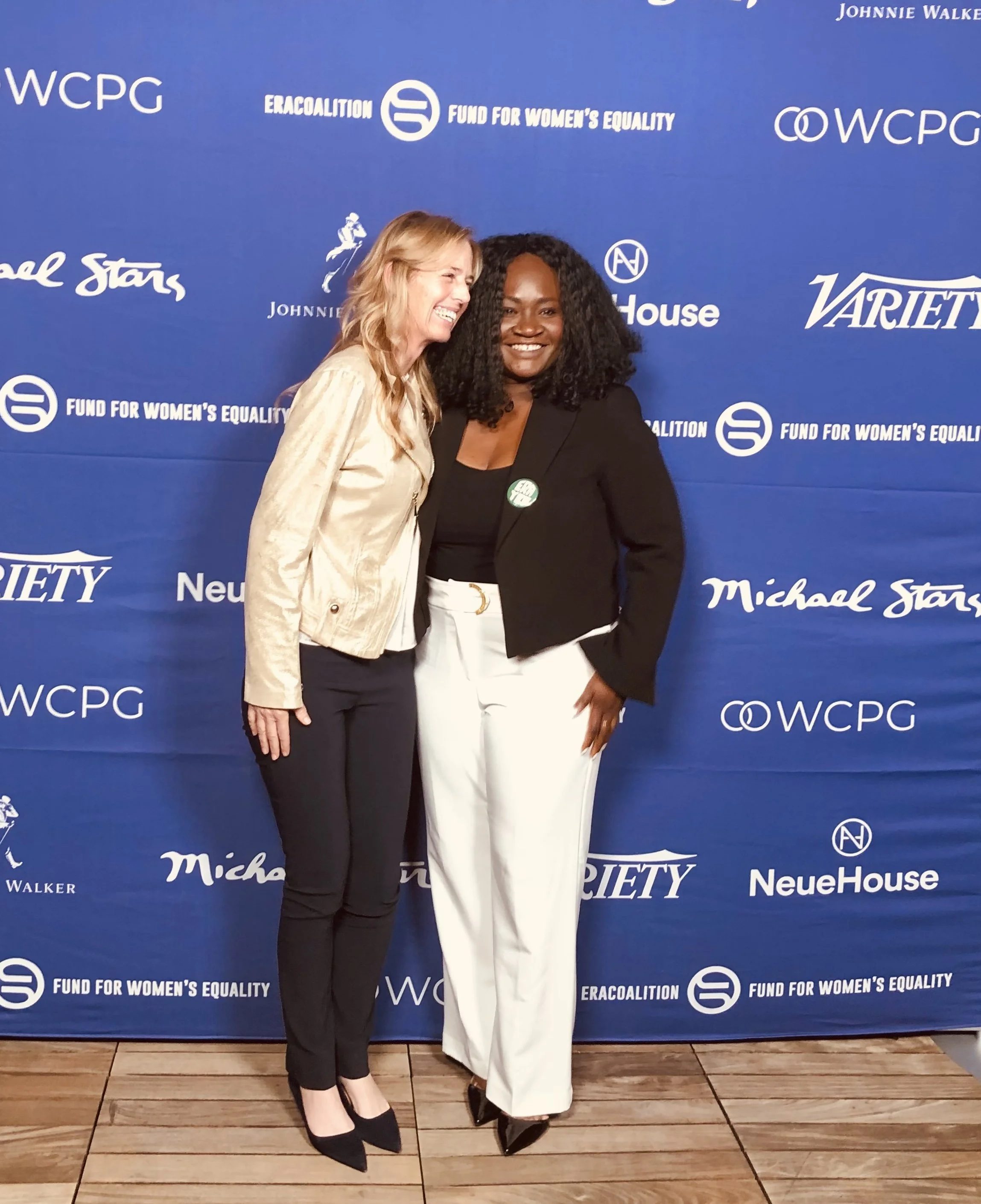 Two women standing together in front of a blue backdrop with multiple logos, smiling and laughing at an event.