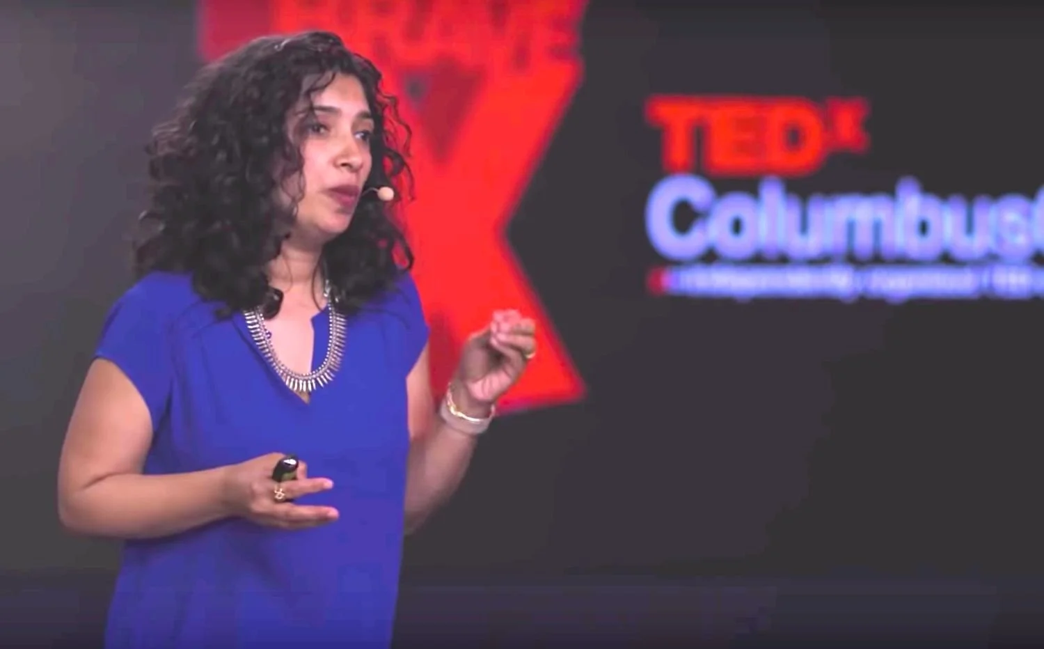 A woman with curly dark hair wearing a blue dress and a silver necklace giving a presentation on a stage with a TedX Columbia backdrop.