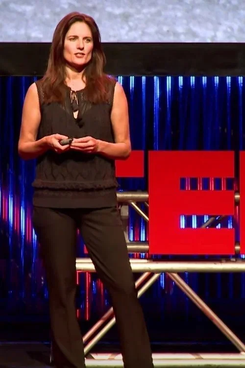 A woman in black attire giving a presentation on a stage with a large red TED logo in the background.