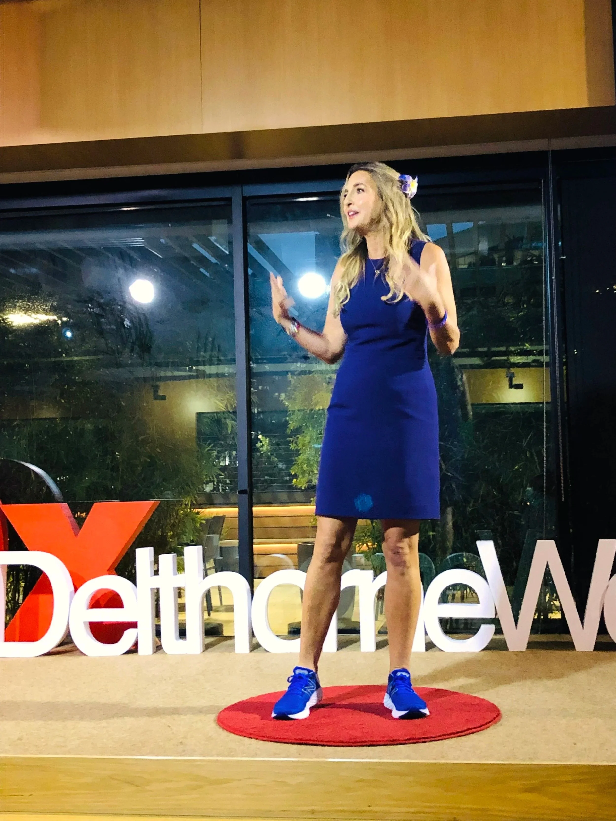 A woman with long blonde hair wearing a blue dress and sneakers speaking on stage at a TEDx event, with the red TEDx circular carpet and a partial view of the TEDx logo on the stage.