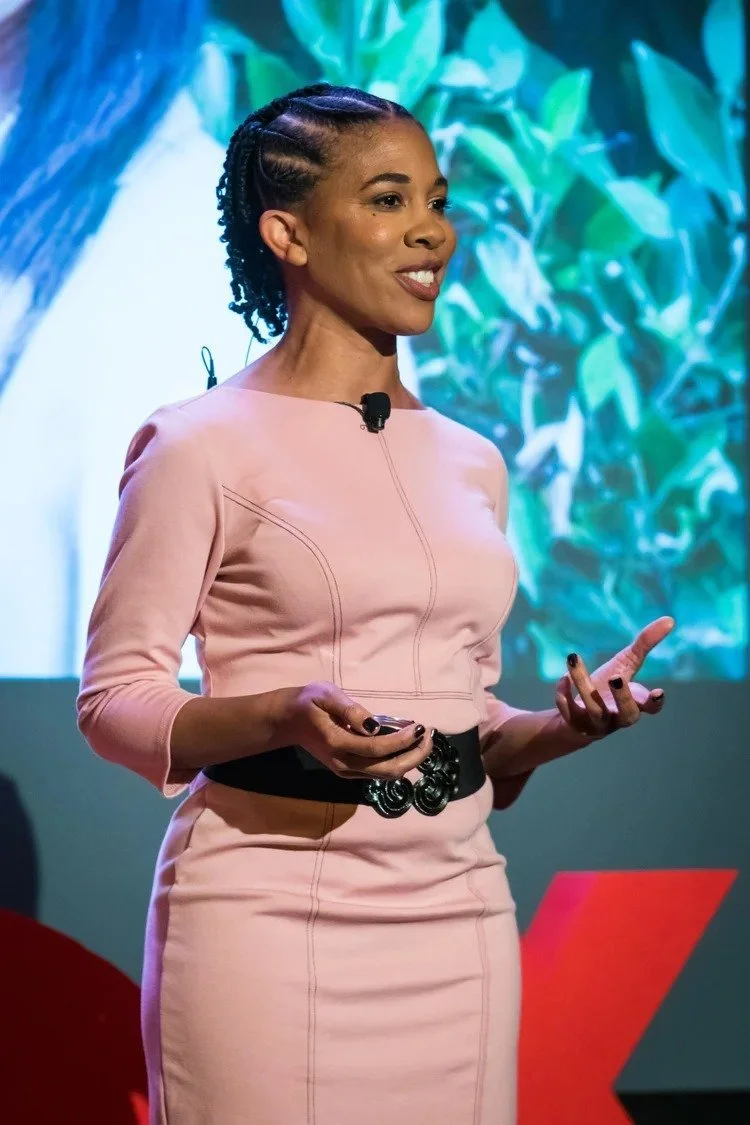 A woman with styled hair and dark skin, wearing a light pink dress with 3/4 length sleeves and a black belt, giving a presentation on stage with a colorful background. She is holding a remote control and gesturing with her left hand.