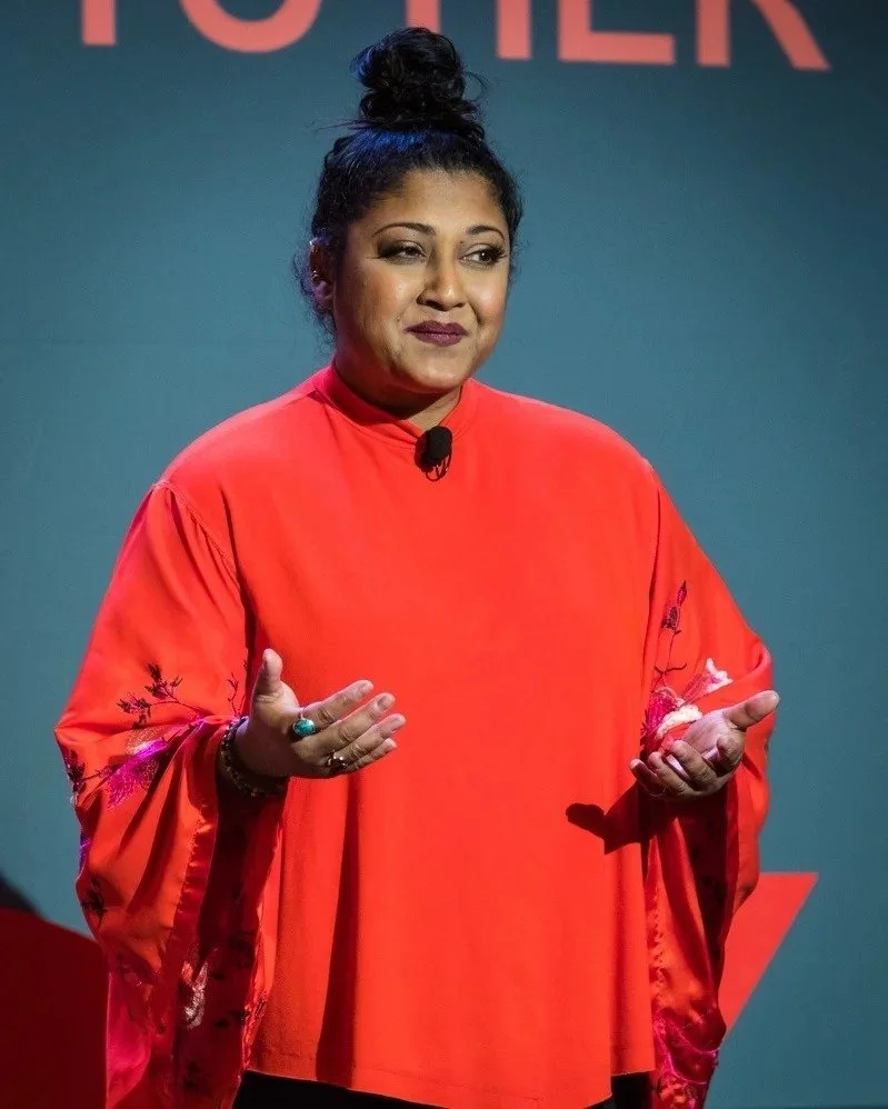 A woman with dark curly hair tied up in a bun, wearing a red blouse with embroidered sleeves, standing in front of a grey background with red text.