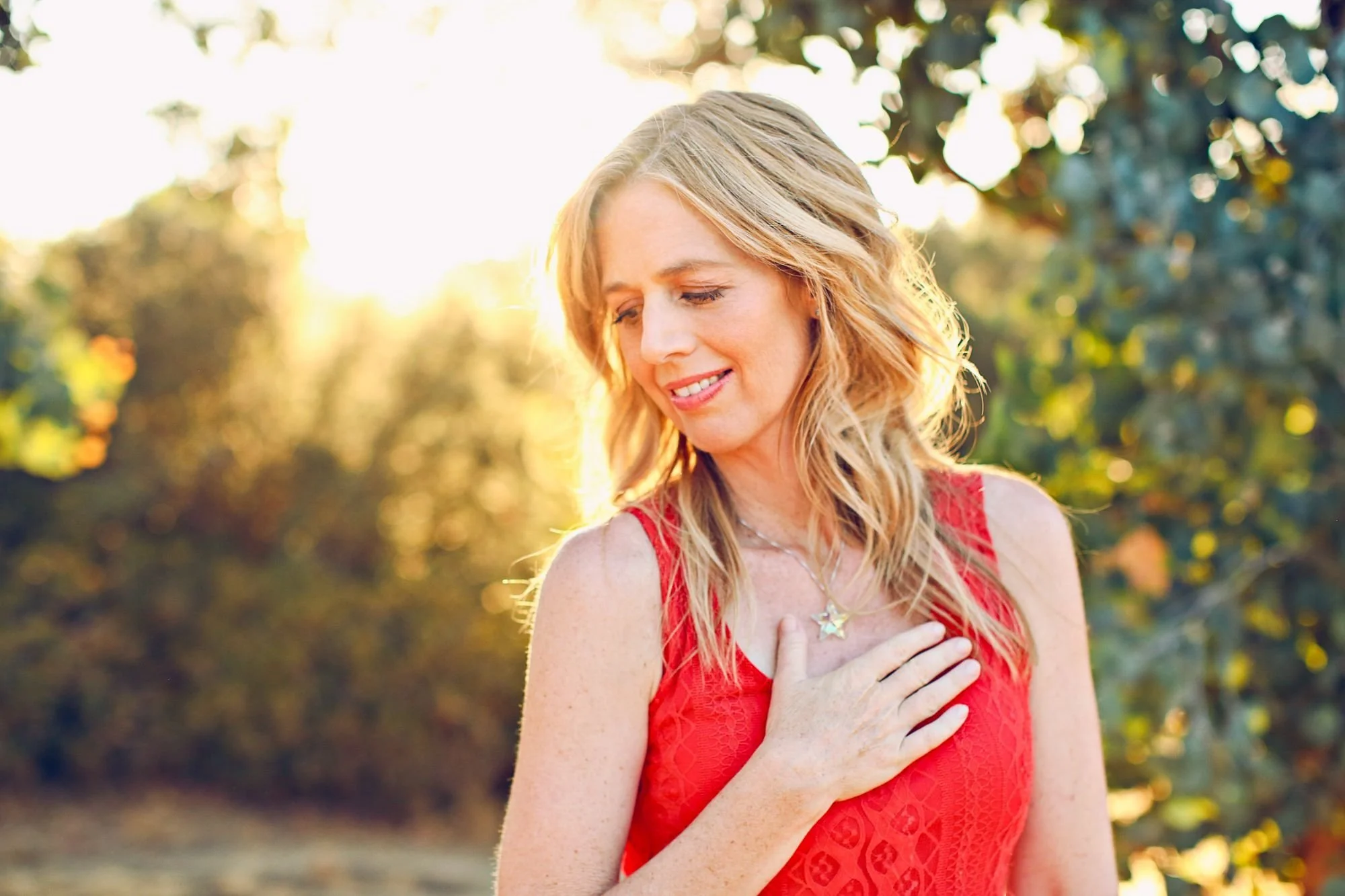 A woman in a red sleeveless dress smiling with her hand on her chest outdoors in sunlight, with trees and foliage in the background.