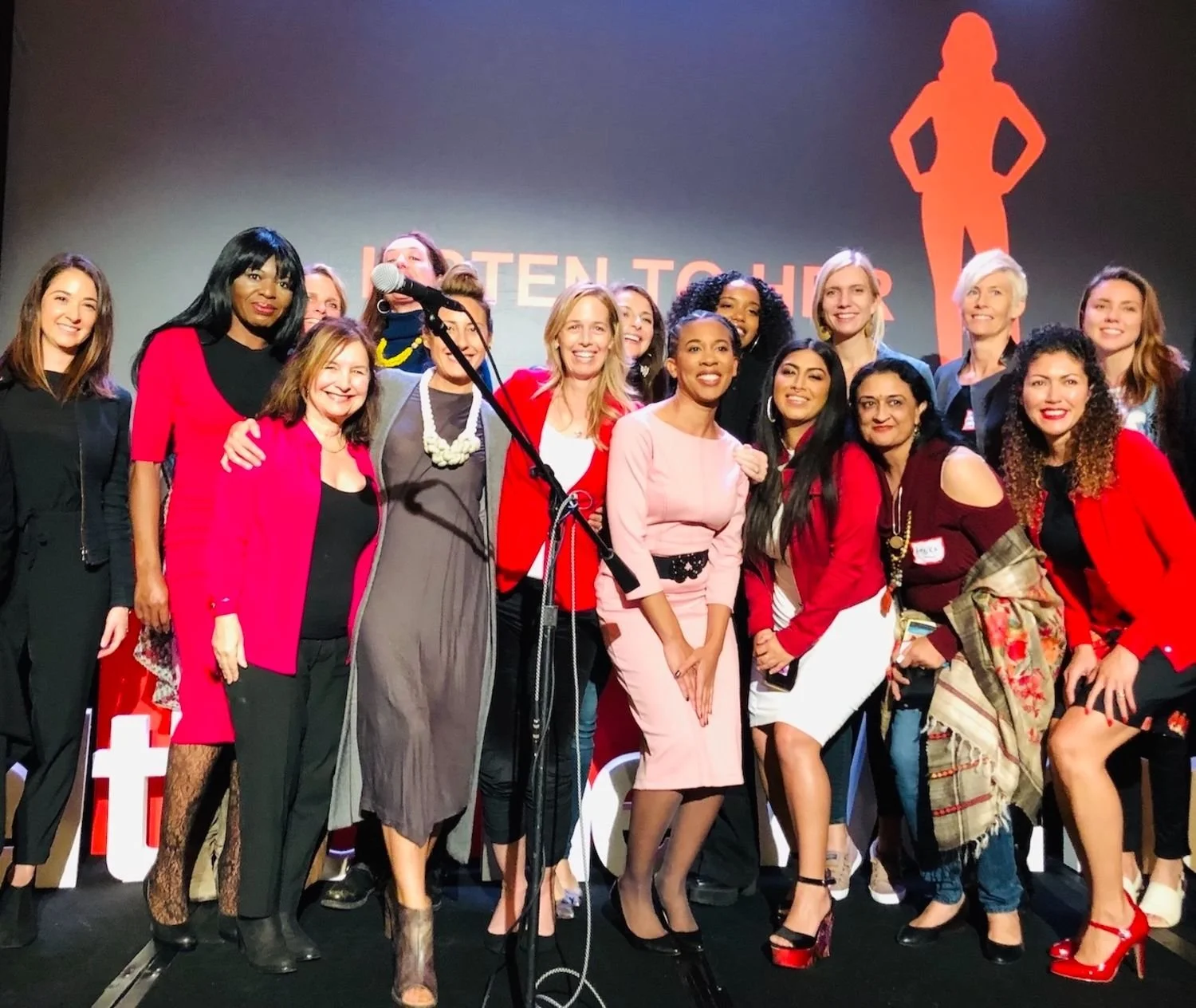 Group of women on stage at an event, with a large female silhouette and the words "Listen to Her" in the background.