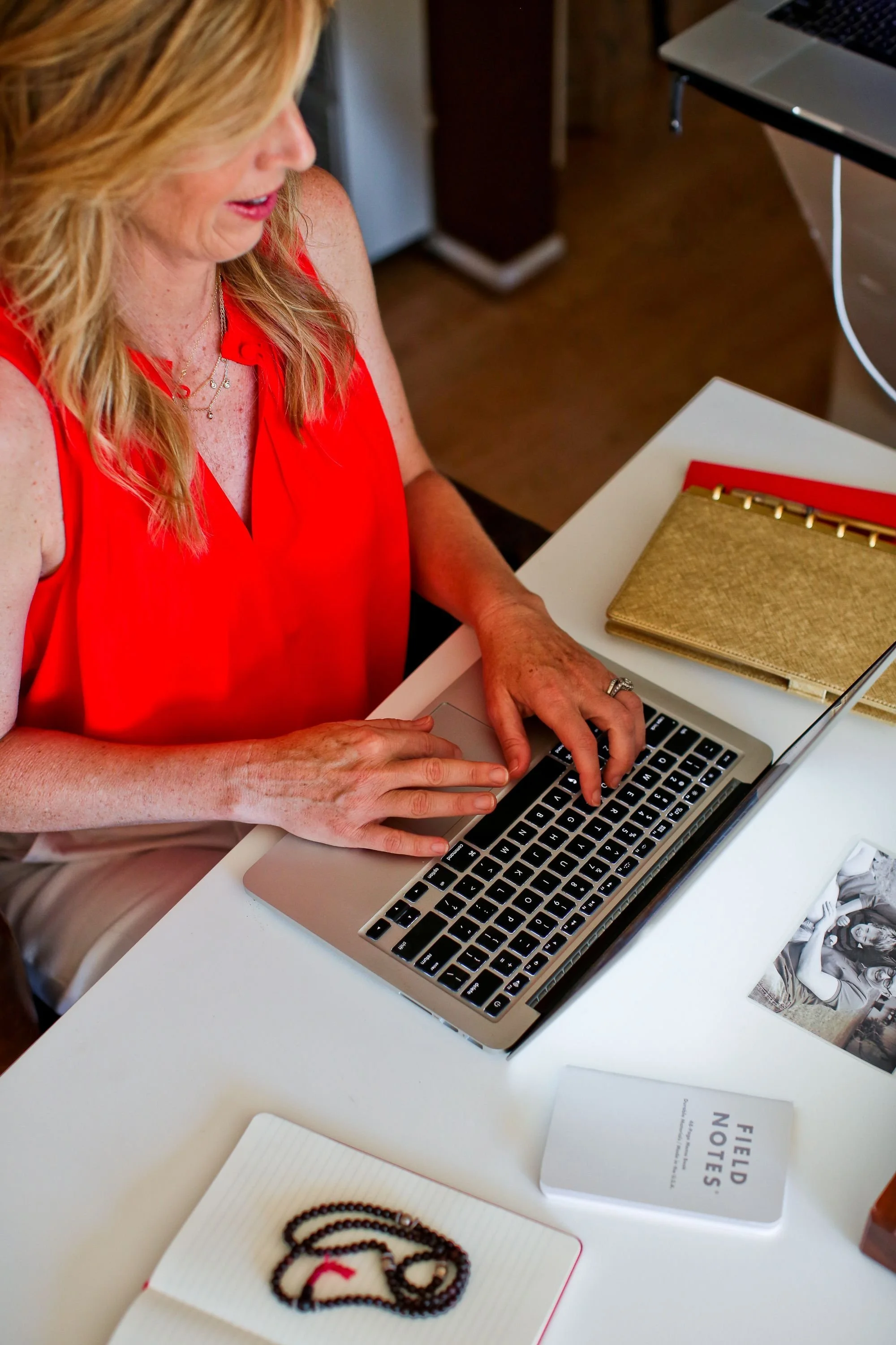 A woman wearing a red sleeveless top typing on a laptop at a white desk with notebooks, a magazine, and a photograph on it.