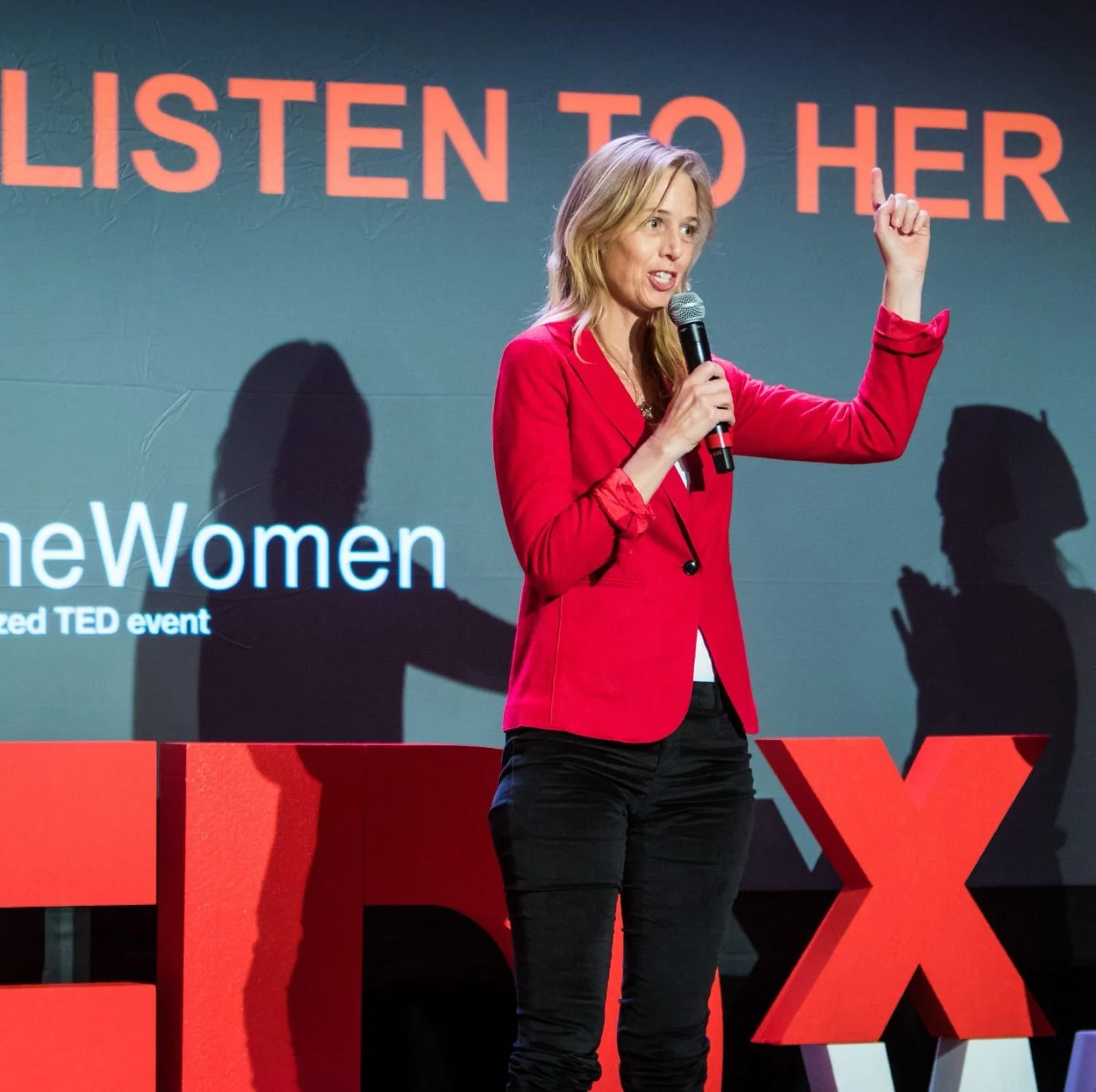 A woman in a red blazer and black pants giving a speech on stage at a TEDx event, with a microphone in her hand. The background features a large screen with the words "LISTEN TO HER" and "Women" and two shadow silhouettes of women.