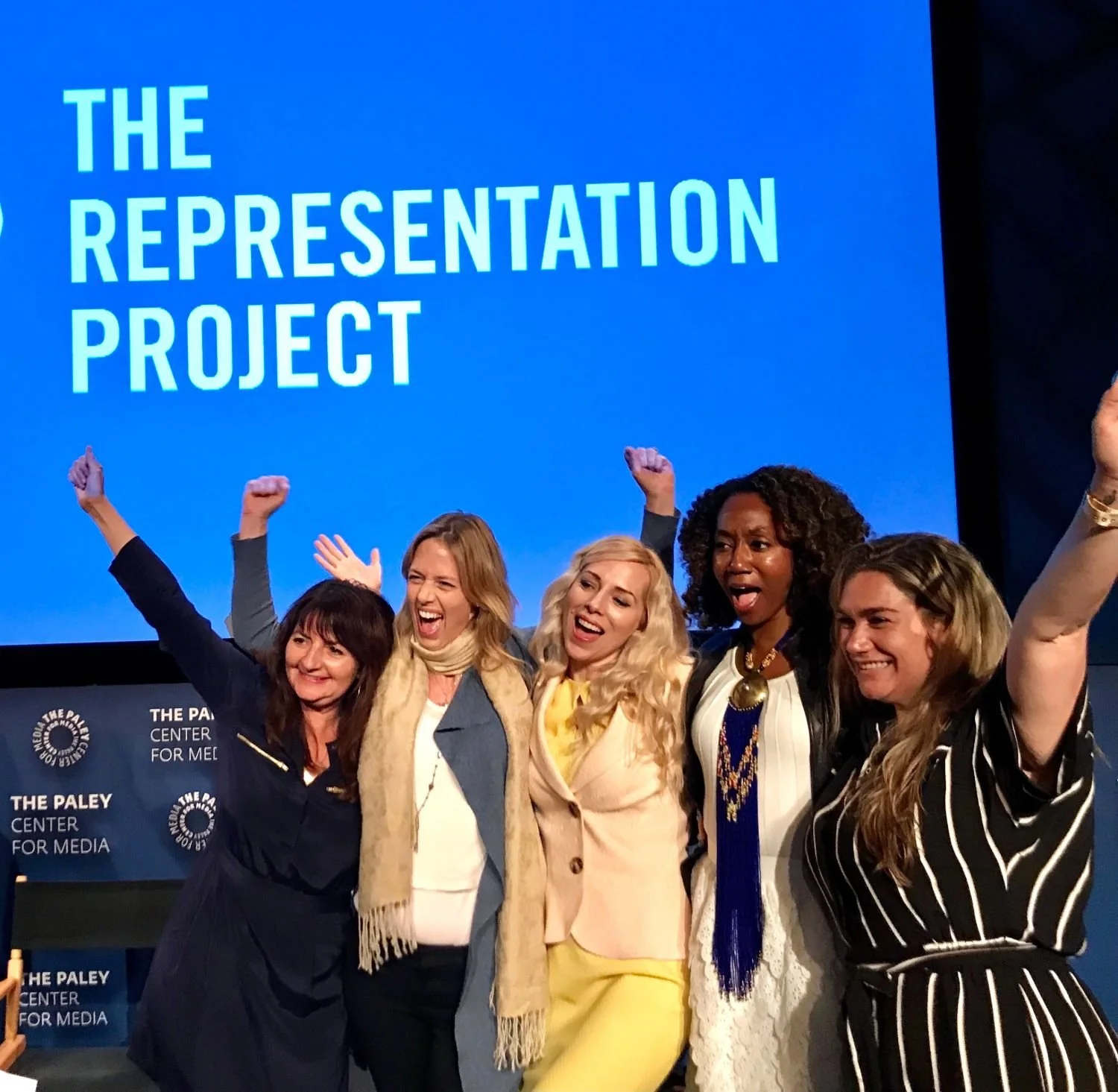 Group of five women on stage, smiling, celebrating, with their arms raised, in front of a blue background with the text 'The Representation Project.'