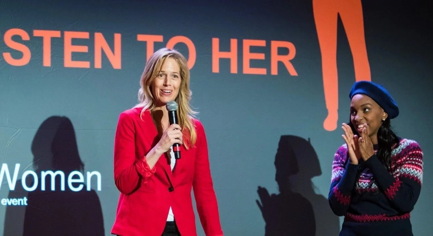 Two women speaking at an event, one holding a microphone, with a large screen behind them displaying partially visible text and a lipstick illustration.